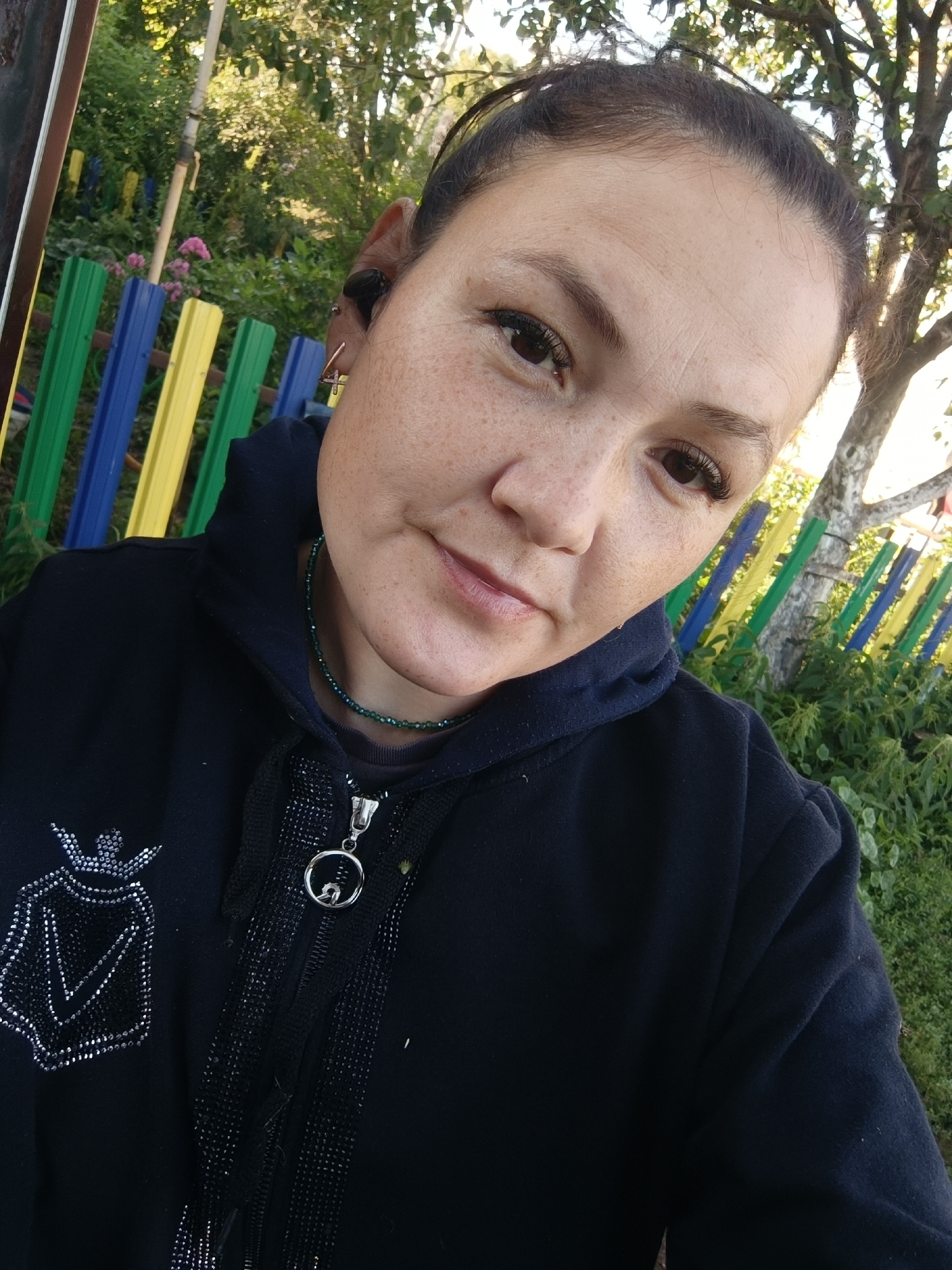 A woman with dark hair pulled back is taking a selfie outdoors. She is wearing a black hooded jacket with a silver crown-like emblem on the left chest and a beaded green necklace. Her right ear is visible, adorned with an earring and an earbud. The background features a colorful fence of vertical wooden planks in blue, yellow, and green, with lush green foliage and trees. The scene suggests a sunny day, possibly in a park or backyard area. There is no indication this image is located in Karsha, Russia.