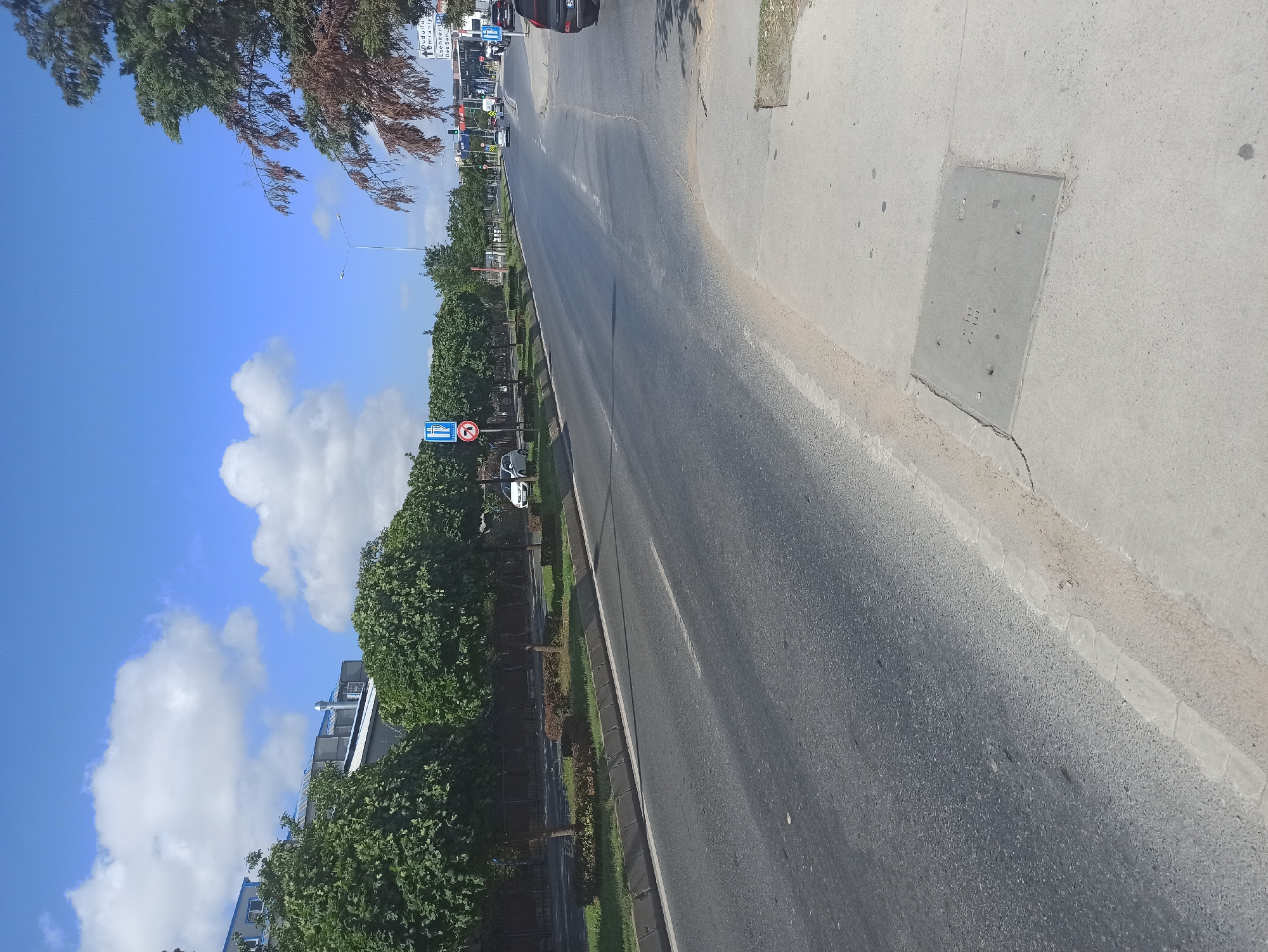 The image depicts a street scene in Ümraniye, Turkey, on a clear day with a bright blue sky and scattered white clouds. The street appears to be a multi-lane road with a concrete sidewalk on the right side. The road surface is asphalt and shows some wear and tear. A row of trees with rounded canopies lines the left side of the road, behind a low fence. Some of these trees appear to be ornamental. On the right side of the road, a few cars are visible, including a dark-colored SUV parked near the curb. Further down the road, there are buildings and more trees. Traffic signs are present, including one with a parking symbol and another indicating a prohibition. A manhole cover is visible on the sidewalk in the foreground. The overall impression is that of an urban environment with moderate traffic.