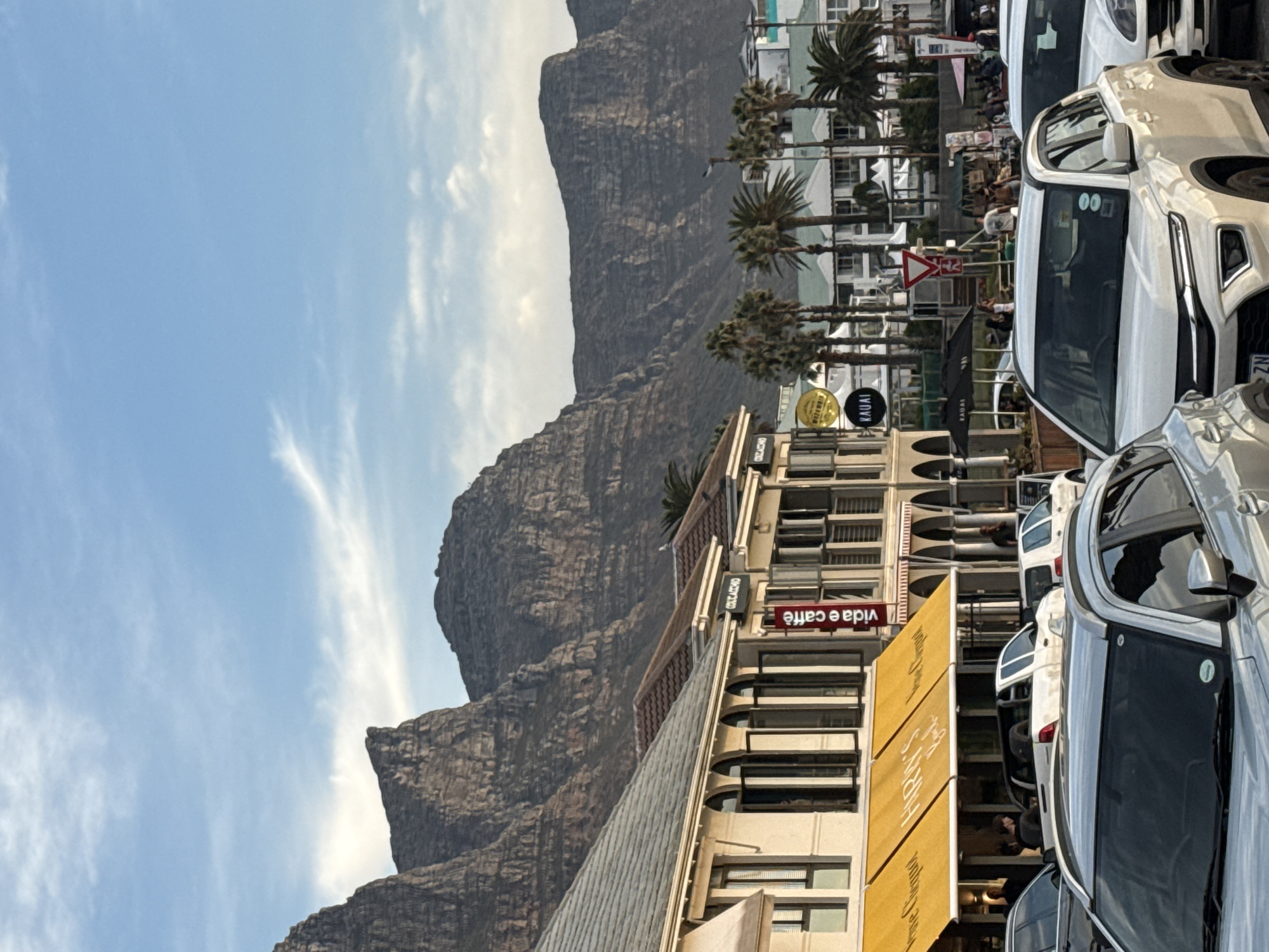 The image shows a street scene in Cape Town, South Africa. In the background, Table Mountain dominates the landscape with a partly cloudy blue sky. In the foreground, buildings line the street, including one with a "vida e caffè" sign. Palm trees are visible along the street. Parked cars fill the right side of the frame. People are visible along the street, though they are not the primary focus. The overall impression is of a busy urban area nestled against a dramatic mountain backdrop.