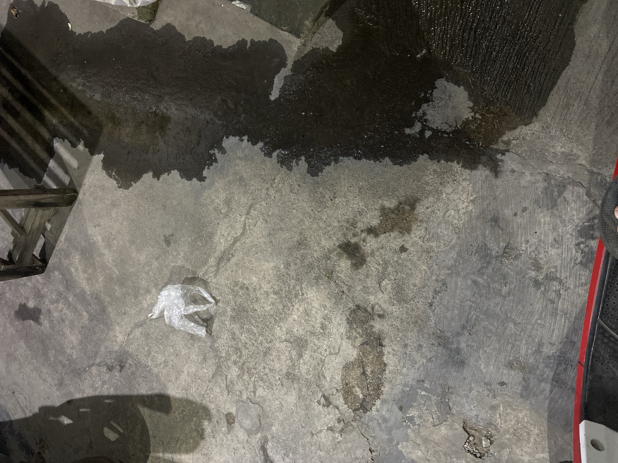 The image shows a wet concrete ground with a white plastic bag on the left side and a large dark puddle on the right. The ground is textured and appears to be outdoors, possibly in a public area given the presence of shadows from what could be a vehicle or furniture. The lighting suggests it might be evening or night. The context of Tulungagung, Indonesia, suggests this is a street or walkway scene in that city.