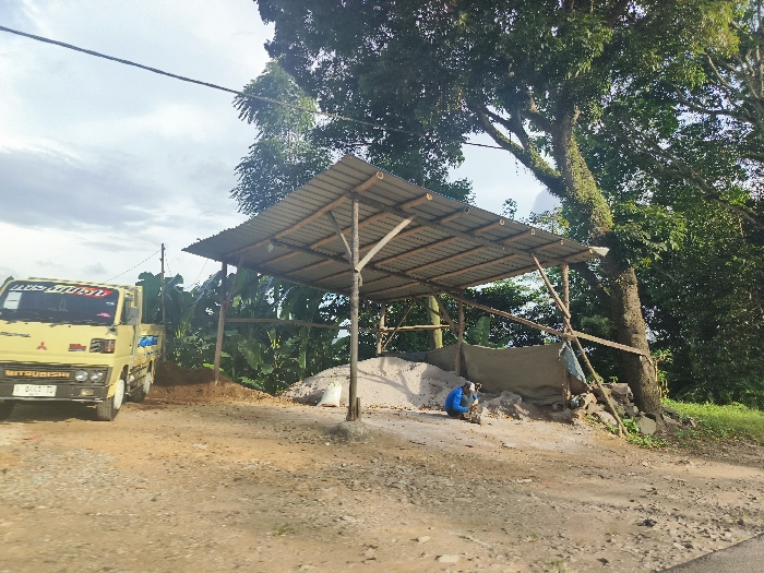 The scene is a dirt lot with a simple metal roof structure. A yellow Mitsubishi truck with the words "LUGSI PUSAT" written on the side is parked next to the structure. There is a pile of dirt/sand to the right of the truck. A man wearing a blue shirt and shorts is seated on the ground to the right of the dirt pile. He appears to be working on something. Behind the man are several rocks and debris. Lush green foliage, including banana trees, fills the background. The sky is blue with a few puffy white clouds. The location is Cikalong, Indonesia, at latitude -6.762 and longitude 107.446.
