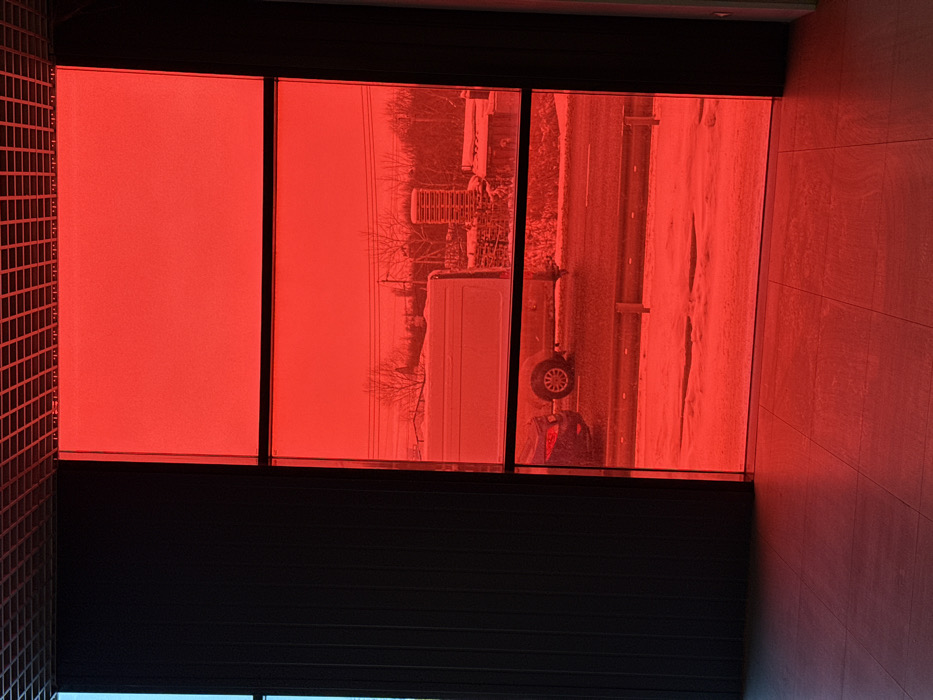 The image depicts an interior perspective looking outward through a large, multi-paned window with a prominent red tint, overlooking an exterior scene in Druzhny, Russia. The interior foreground features a light-colored, tiled floor reflecting the red hue of the window. A grid-patterned ceiling is visible at the top, and dark vertical panels or blinds are present on the left, partially obscuring a narrow, faintly blue-tinted window section.
The exterior scene, viewed through the red tint, indicates cold weather with snow covering the ground. A cleared, dark road traverses horizontally across the mid-ground, flanked by a snow-covered verge and small, dark barriers. A light-colored panel van is positioned on the right side of the road, facing left, its side visible. Behind it and slightly to the left, the rear section of a dark-colored sedan is partially visible. In the background, beyond the road and snow, bare deciduous trees and utility poles with power lines are present. A light-colored, possibly cylindrical stack of objects or an industrial structure is discernible in the far right background. No people are visible in the scene.