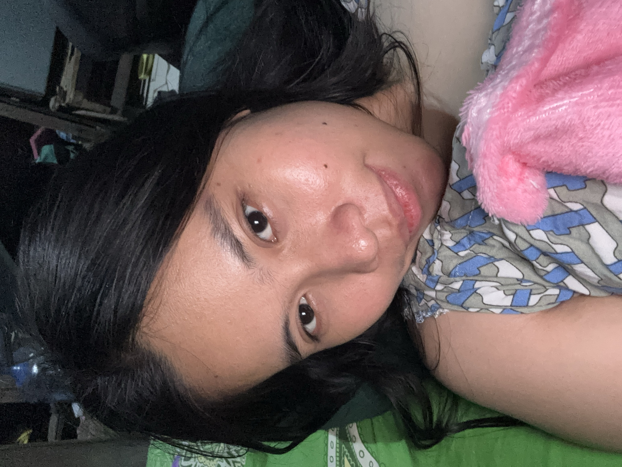 A close-up, high-angle selfie captures a young woman with dark hair lying on a bed in East Jakarta, Indonesia. Her face is turned towards the camera, and she is looking directly into the lens with a slight smile. She is wearing a patterned top and a pink fluffy blanket is visible. The background is blurred, but appears to be a bedroom with some furniture and objects. The lighting is dim and appears to be from an artificial source.