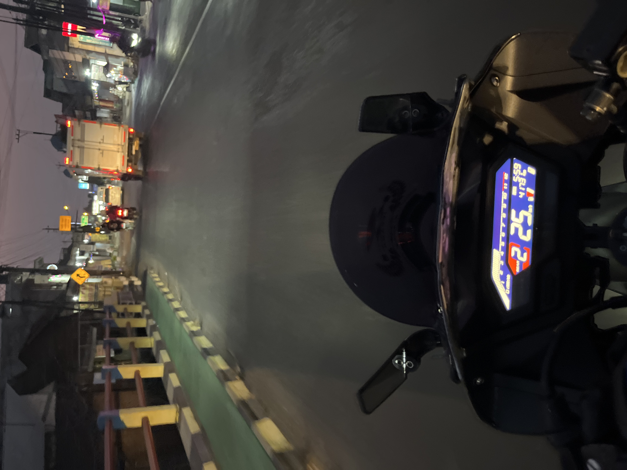 The image is taken from the perspective of someone riding a motorcycle at dusk on a street in Kalinyamatan, Indonesia. The motorcycle's digital dashboard is visible in the foreground, displaying a speed of 25 km/h and the time as 5:59. The road is paved, with yellow and black striped barriers and a green path on the left side, suggesting a pedestrian or bicycle lane. Buildings with illuminated signs line the street, indicating commercial establishments. Traffic is present, with several motorcycles and a truck visible. The sky is a dusky purple, suggesting the transition from day to night. The overall scene depicts a typical streetscape in a Indonesian town during the evening hours, with active road traffic and illuminated businesses.