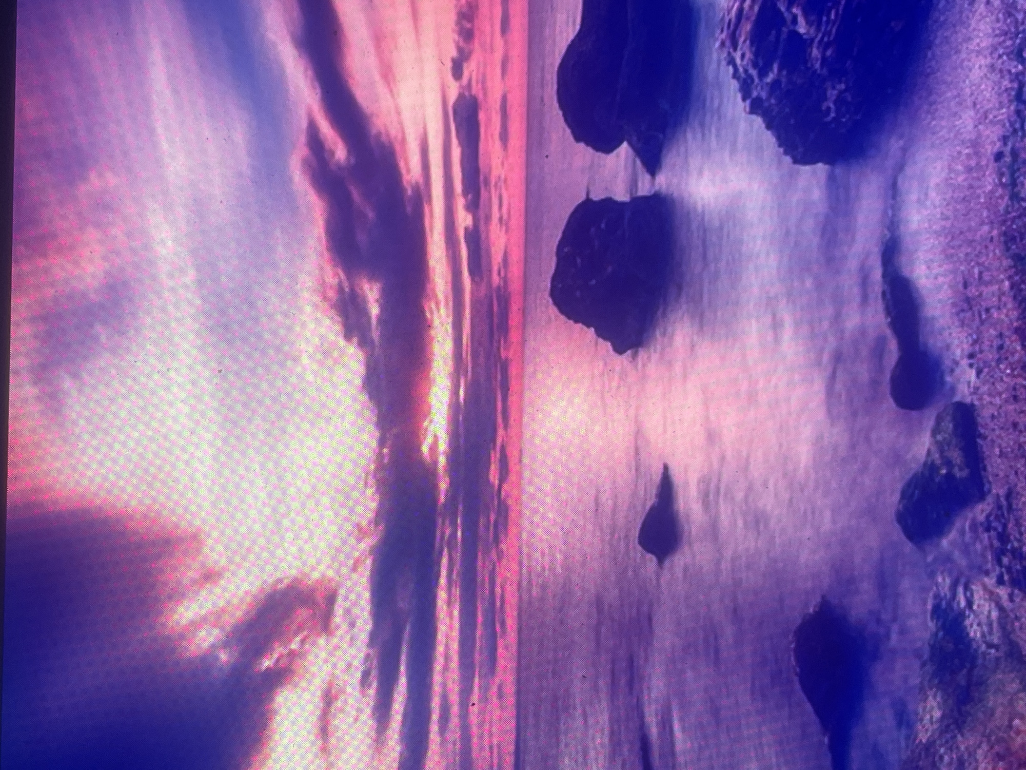 The image captures a serene coastal landscape at either sunrise or sunset, located in the city of Zelenyi Hai, Ukraine. The upper portion of the scene is dominated by a sky displaying vibrant pink and purple hues, with light rays emanating from a bright spot on the horizon, indicative of the sun's position. Clouds, varying in density and shape, are scattered across the sky, reflecting the intense colors.
Below the horizon, the sea mirrors the sky's palette, appearing in shades of pink and purple. The water's surface is smooth and exhibits a slightly blurred texture, suggesting a long-exposure photograph that softens movement. Several dark, irregular rock formations are present within the water. Two sizable rocks are prominently located in the mid-right, partially submerged. A smaller, solitary rock is visible in the mid-left area. The immediate foreground reveals a rocky shoreline composed of smaller stones and pebbles, with larger dark rocks interspersed along the water's edge.
No individuals, animals, or man-made structures are present in the scene. The overall image features a visible grid pattern across its entirety, consistent with a photograph captured from a digital screen.