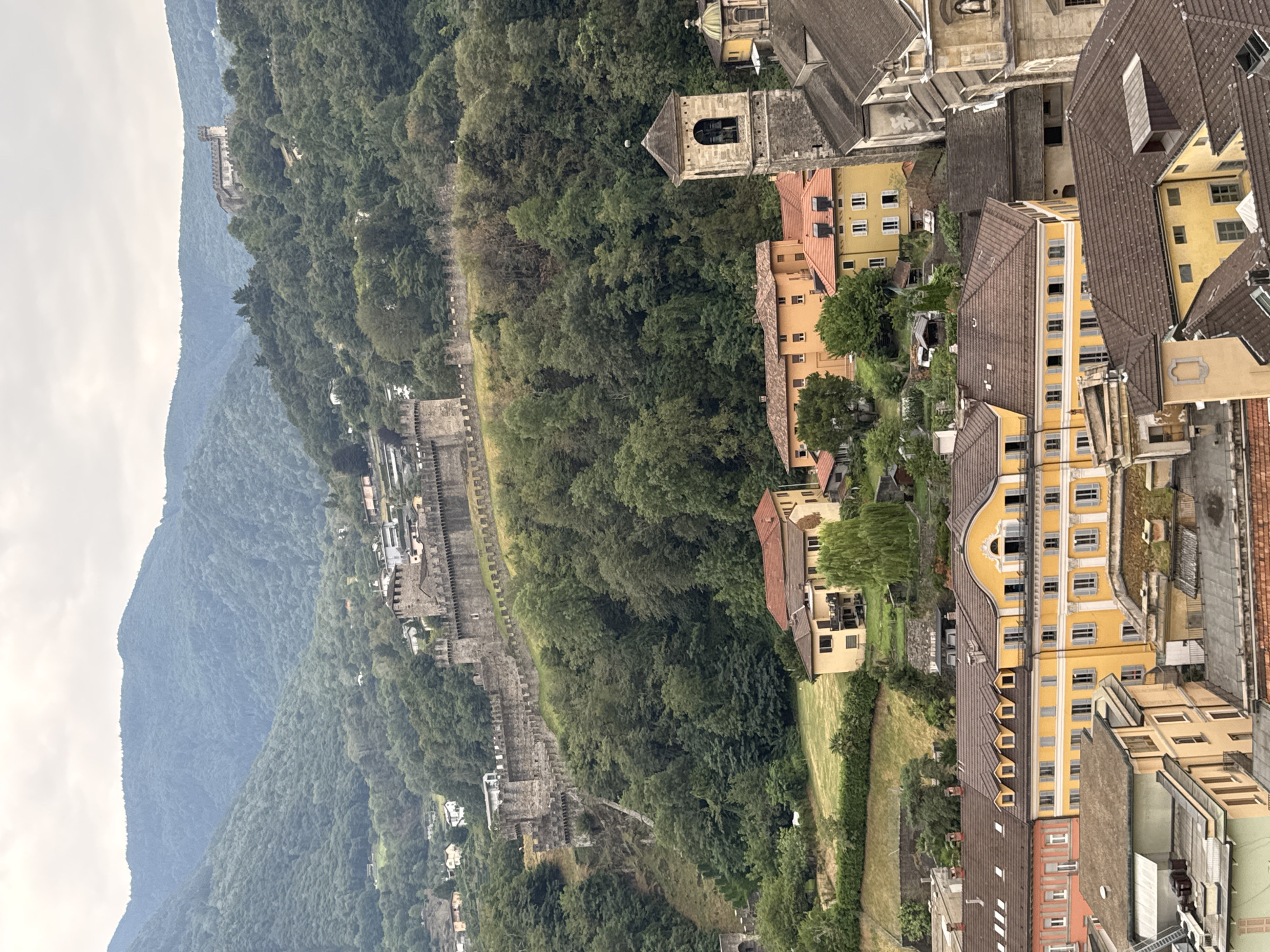 This is a high-angle shot of the city of Bellinzona, Switzerland, focusing on its historic castles and the surrounding landscape. In the foreground, a cluster of buildings with red tile roofs and yellow facades are visible, some with architectural details like dormer windows and awnings. A prominent bell tower with a stone base and a tiled roof stands out among the buildings. Further up the hillside, a long, fortified stone wall snakes across the landscape, connecting to a castle structure. The background is dominated by lush green mountains covered in dense trees, with a hazy sky overhead. A more distant castle, Castello Montebello, can be seen on a higher peak to the left. The scene depicts a blend of urban architecture and natural scenery, with a strong emphasis on the medieval fortifications.
