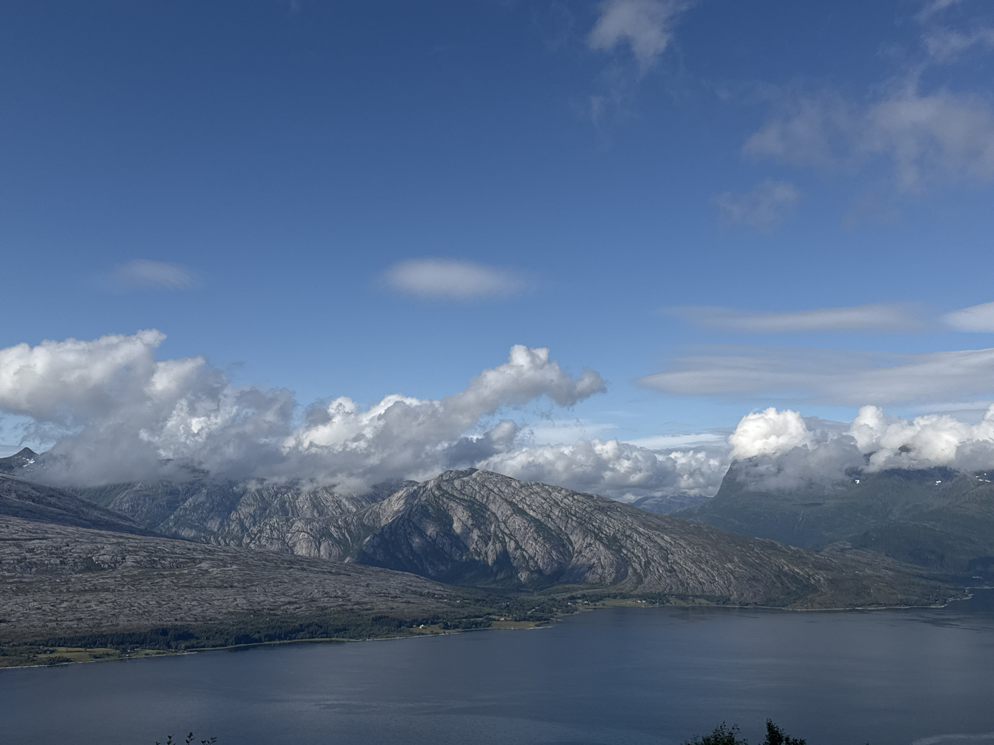 The image depicts a vast, serene landscape of mountains and a body of water, likely a fjord, under a bright blue sky dotted with scattered white clouds. The foreground is dominated by the dark blue water, reflecting the sky. Beyond the water, a textured, rocky mountainside rises, its surface appearing weathered and uneven. Further in the distance, more rugged peaks emerge, some partially obscured by white, puffy clouds that are clinging to their slopes. A strip of green forest separates the rocky terrain from the water's edge, with a few scattered structures suggesting a small settlement. The overall impression is one of natural grandeur and tranquility. The geographical location is identified as Flostrand, Norway.