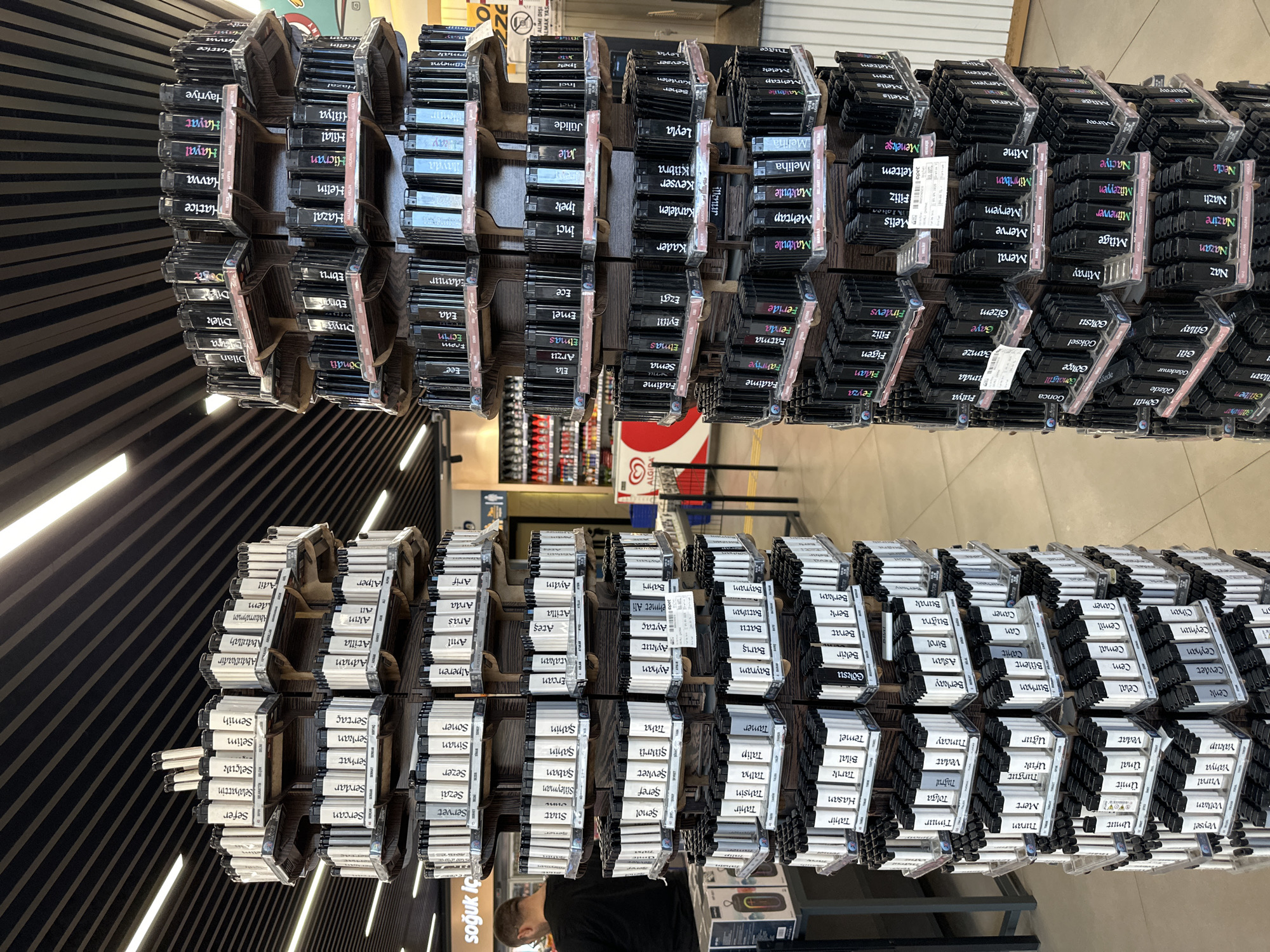 The image shows shelves of personalized lighters with names on them. They are arranged in rows. The lighters are black and have labels with white writing. In the bottom-left corner, a person is partially visible, wearing a dark shirt and looking down, likely browsing the products. The background includes store shelves with various items, and ceiling details. The text "soğuk İç" is visible, indicating a food and beverage section. Based on the OCR text mentioning "ALGI," which could refer to the Algida brand (ice cream), there are Algida freezers in the background. The text "ALMAK YAS" implies a restriction, and "ALIMI DISE" seems to be a sales-related message. It is located in a store, presumably in the city of Gaziosmanpaşa, Turkey.