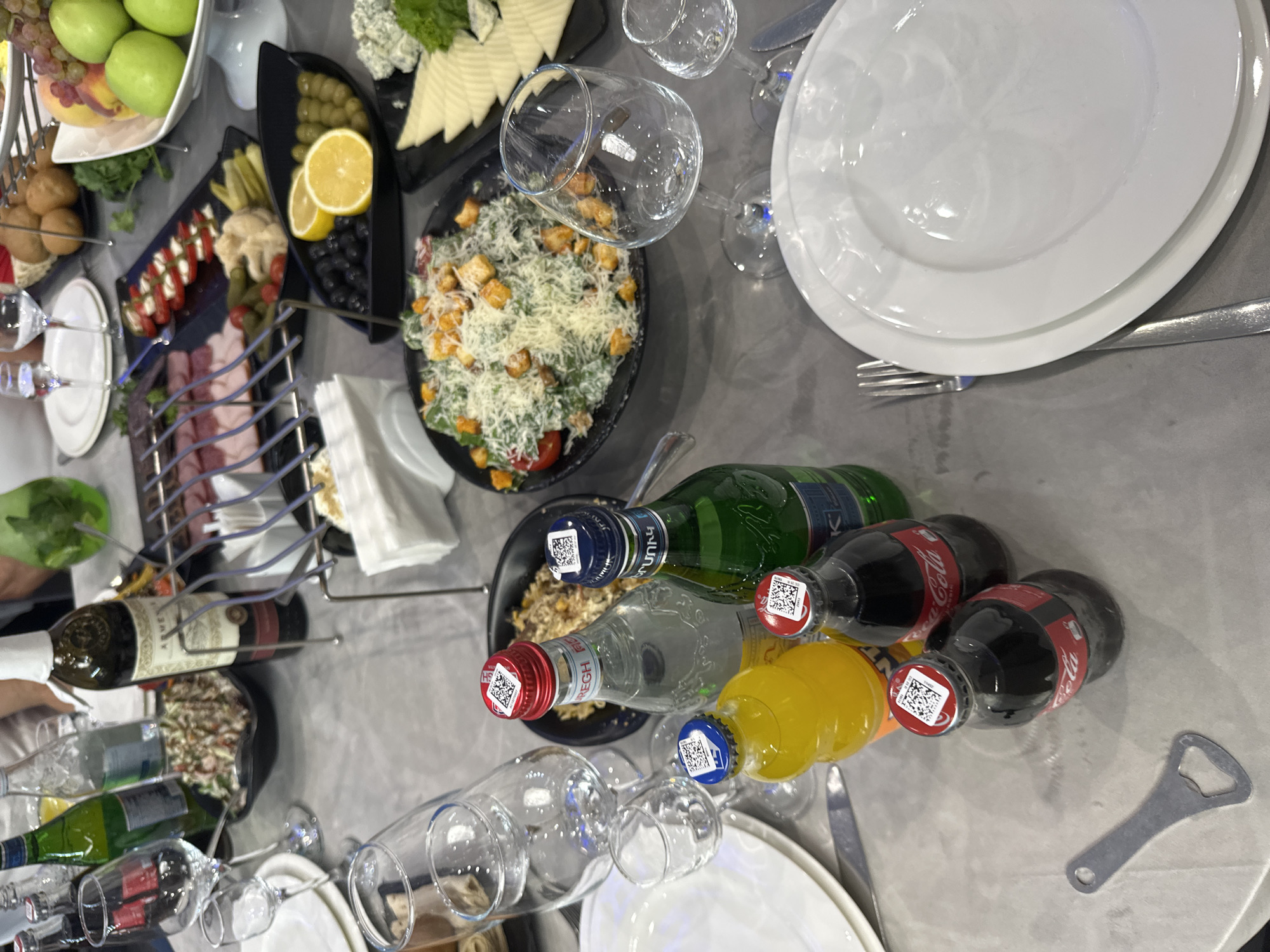 The image depicts a formal dinner setting in Parakar, Armenia, featuring a banquet table laden with food and drinks. A variety of dishes are visible, including a salad, sliced meats, pickled vegetables, olives, lemons, and a cheese platter. Several bottles of beverages, including Coca-Cola and other soft drinks, are arranged on the table, alongside wine glasses and water glasses. In the background, a person can be seen holding a glass, suggesting an ongoing meal or celebration. The overall ambiance appears to be one of a festive gathering, possibly a wedding or a special occasion. The table is set with white plates and silverware, and the environment suggests an indoor dining area.