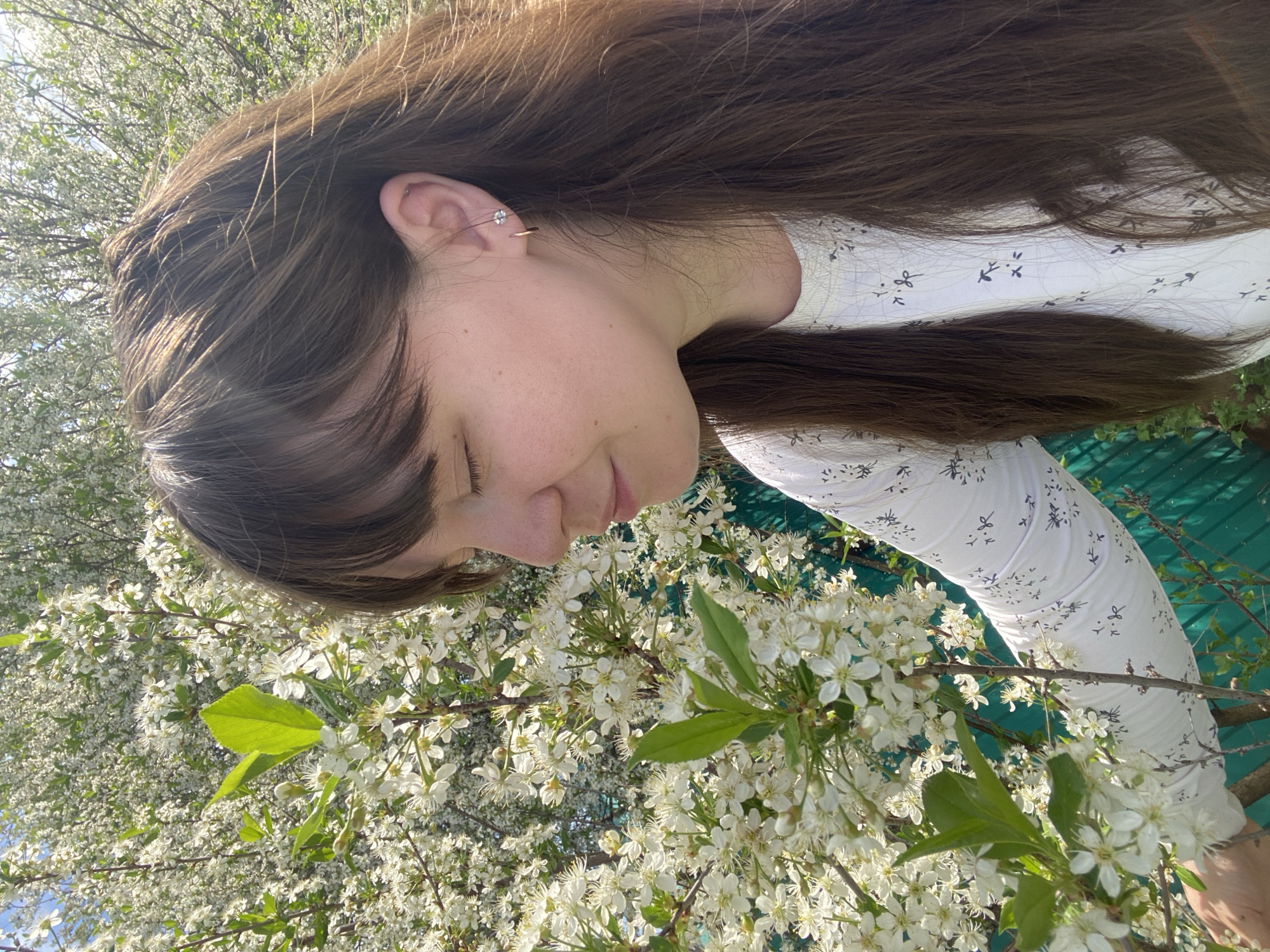 A woman with long brown hair and a white shirt with a small floral pattern stands next to a blooming tree with white flowers. She is facing downwards, her eyes are closed, and she has a slight smile. Her left ear is visible and features a small earring. The background includes more blooming branches and some green structures. Location: Cannot be determined to be Votkinsk, Russia based on visual information.