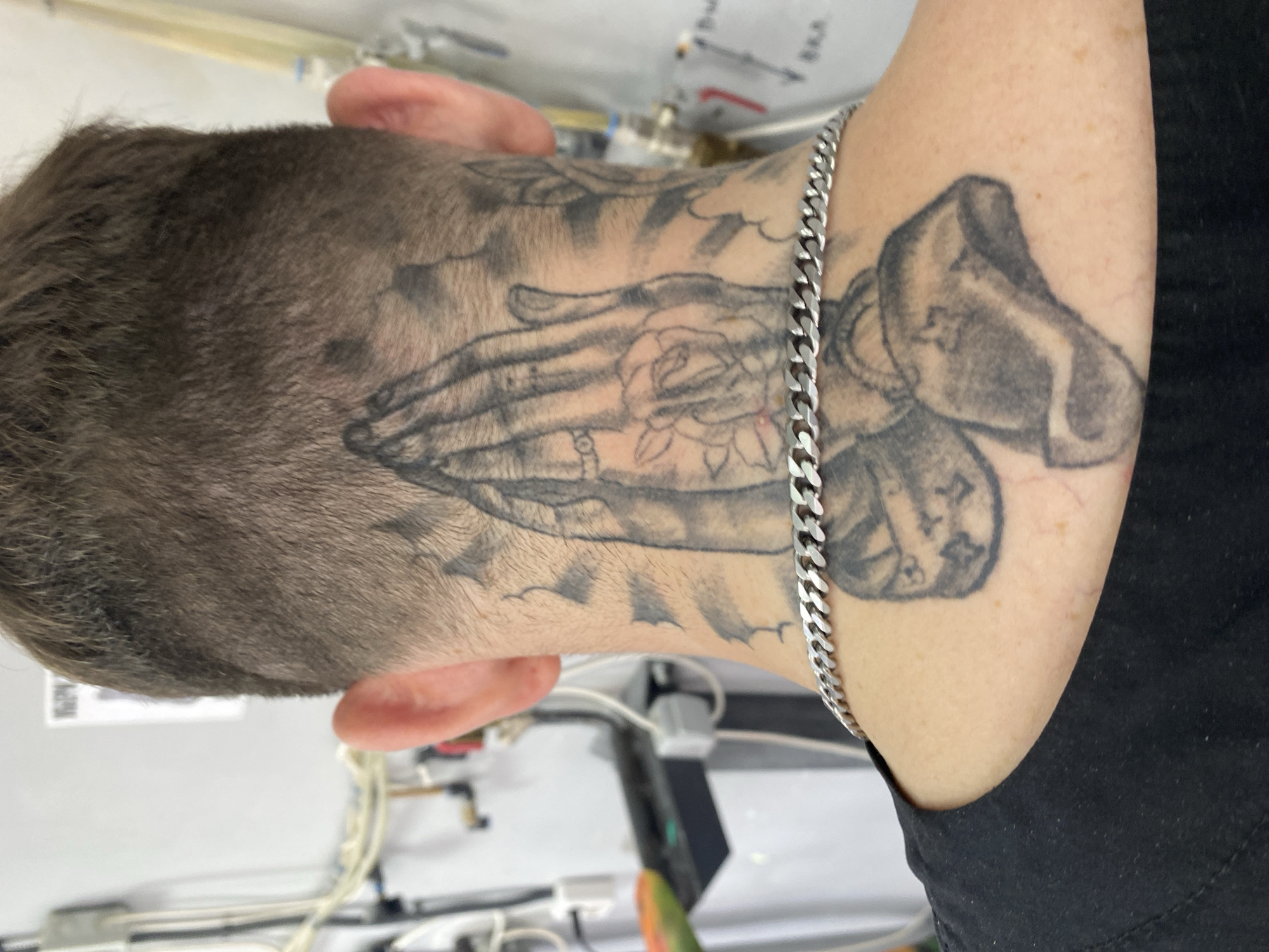 The image shows the back of a person's head and neck, with a tattoo of praying hands and a rose on their neck. A silver chain necklace is visible around their neck. The background appears to be indoors, with some pipes and machinery visible. The individual's hair is short and styled in a fade. The image is likely taken in Vladimir, Russia, based on the prompt's context.