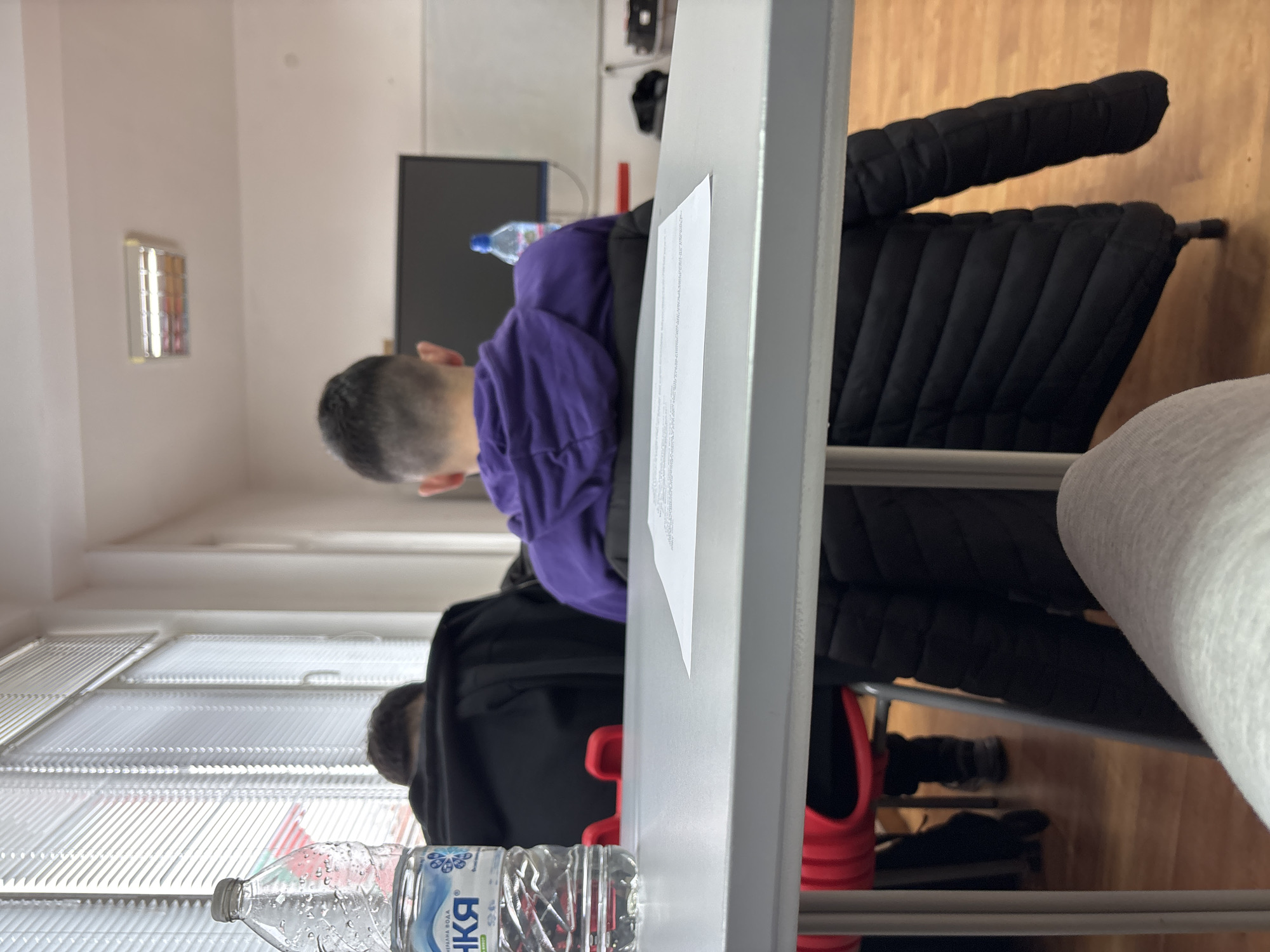 A slightly over-exposed, low-angle, interior shot shows a room with a person wearing a purple hooded sweatshirt, with their back to the camera. There are a few other people present but they are not easily identifiable. A black puffy jacket is draped over a chair. A clear plastic bottle, labeled "NIKA" in Cyrillic characters, sits on a light grey table in the foreground. There is a partially visible stack of papers resting on the table. Windows are present with white blinds. Wood floors are visible in the background. A flat-screen television is visible mounted on the wall. The room contains multiple tables. The exact location in Zlatograd, Bulgaria, cannot be determined from the information presented.