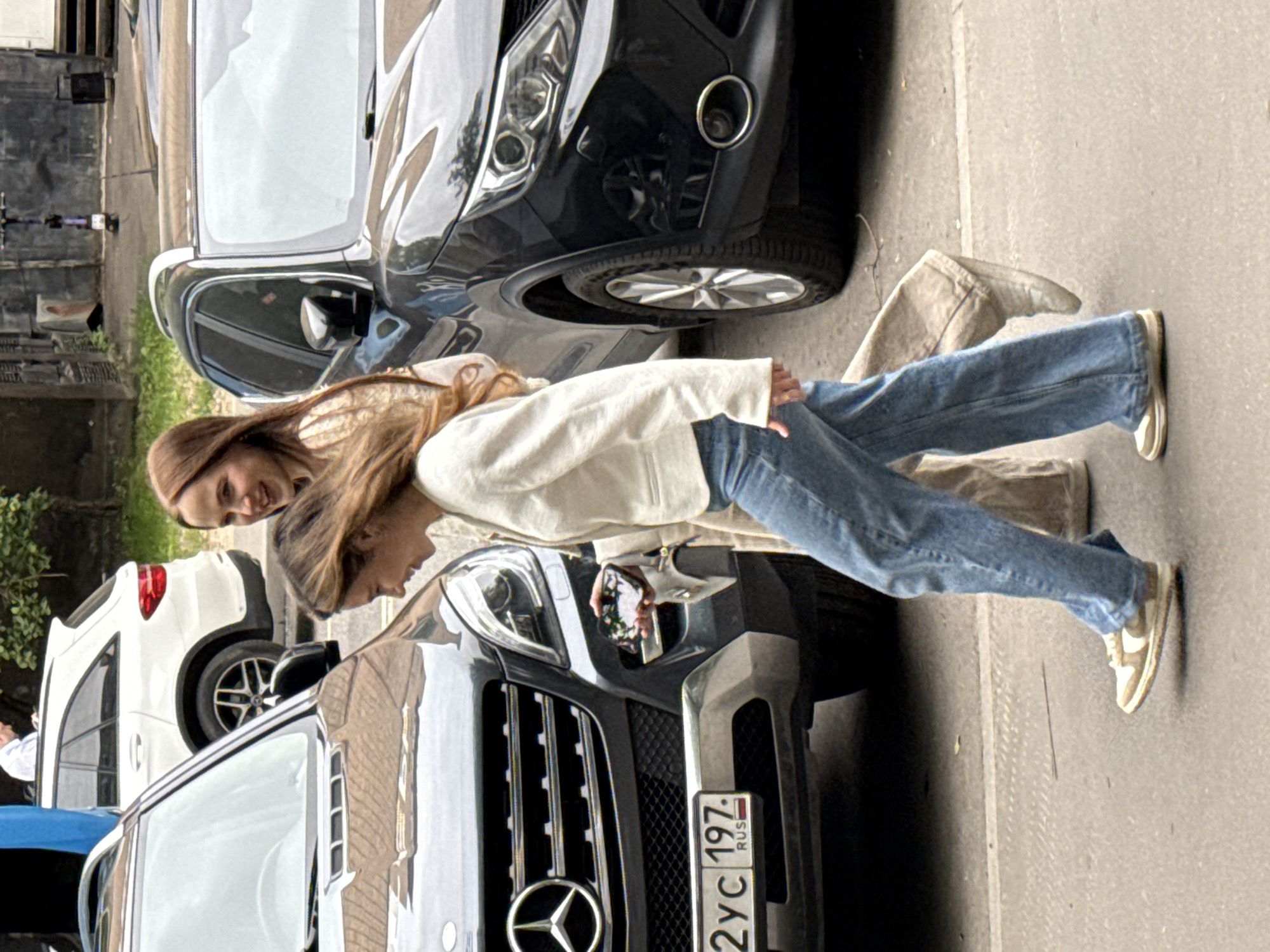 Two young women are depicted in an outdoor urban setting, surrounded by parked cars. One woman, with light brown hair and a light-colored top, stands upright and smiles directly at the camera. The other woman, also with light brown hair and wearing a white jacket over blue jeans, leans forward, looking down at the front of a dark SUV, possibly holding a phone. She is wearing light-colored sneakers.
The prominent vehicle in the foreground is a dark-colored Mercedes-Benz SUV, identifiable by its distinctive front grille and emblem. Its license plate, "2YC 197 RUS," is clearly visible on the front bumper. The "RUS" indicates Russia, and "197" is a code for Moscow, confirming the location context as Moscow, Russia. Behind the women, another dark car is visible, and further back, a white SUV is parked. The ground is paved, suggesting a street or parking area. The lighting indicates it is daytime, possibly overcast, given the soft, diffused light without harsh shadows. The overall mood appears casual and relaxed.