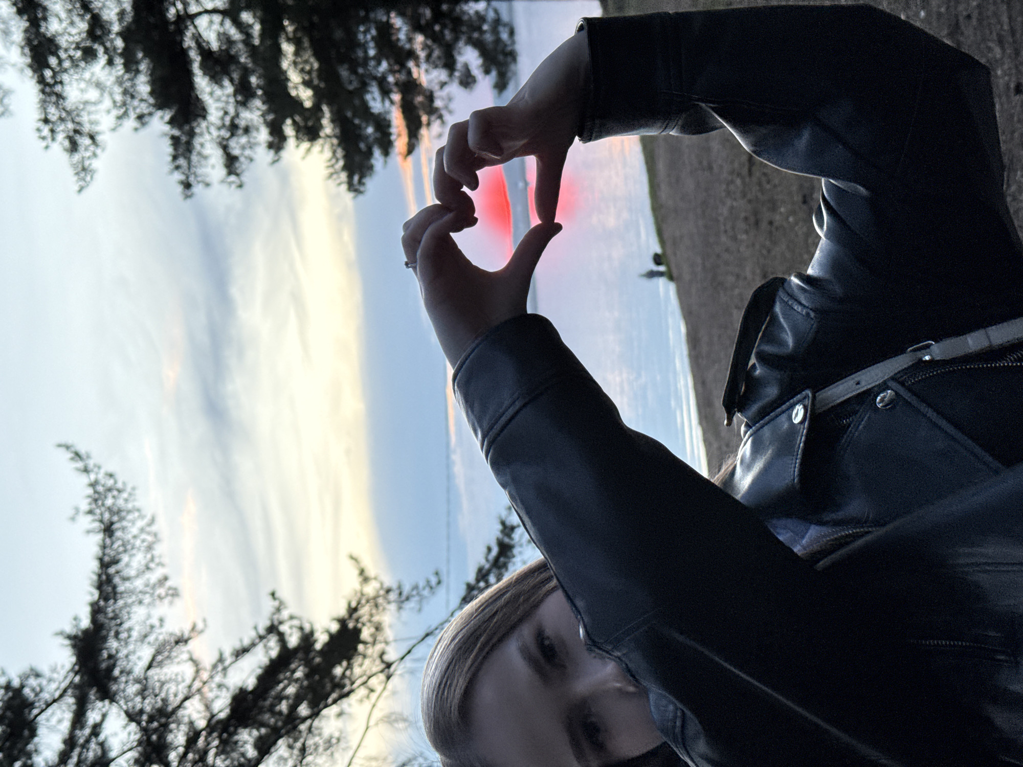 A rotated close-up shows a fair-skinned person in a black leather jacket making a heart shape with their hands against a backdrop of water and sky. The sky transitions from light blue at the top to yellow and pink at the horizon, reflecting red light onto the water's surface. A treeline is visible along the edge of the image. The location is Chaikovsky, Russia.