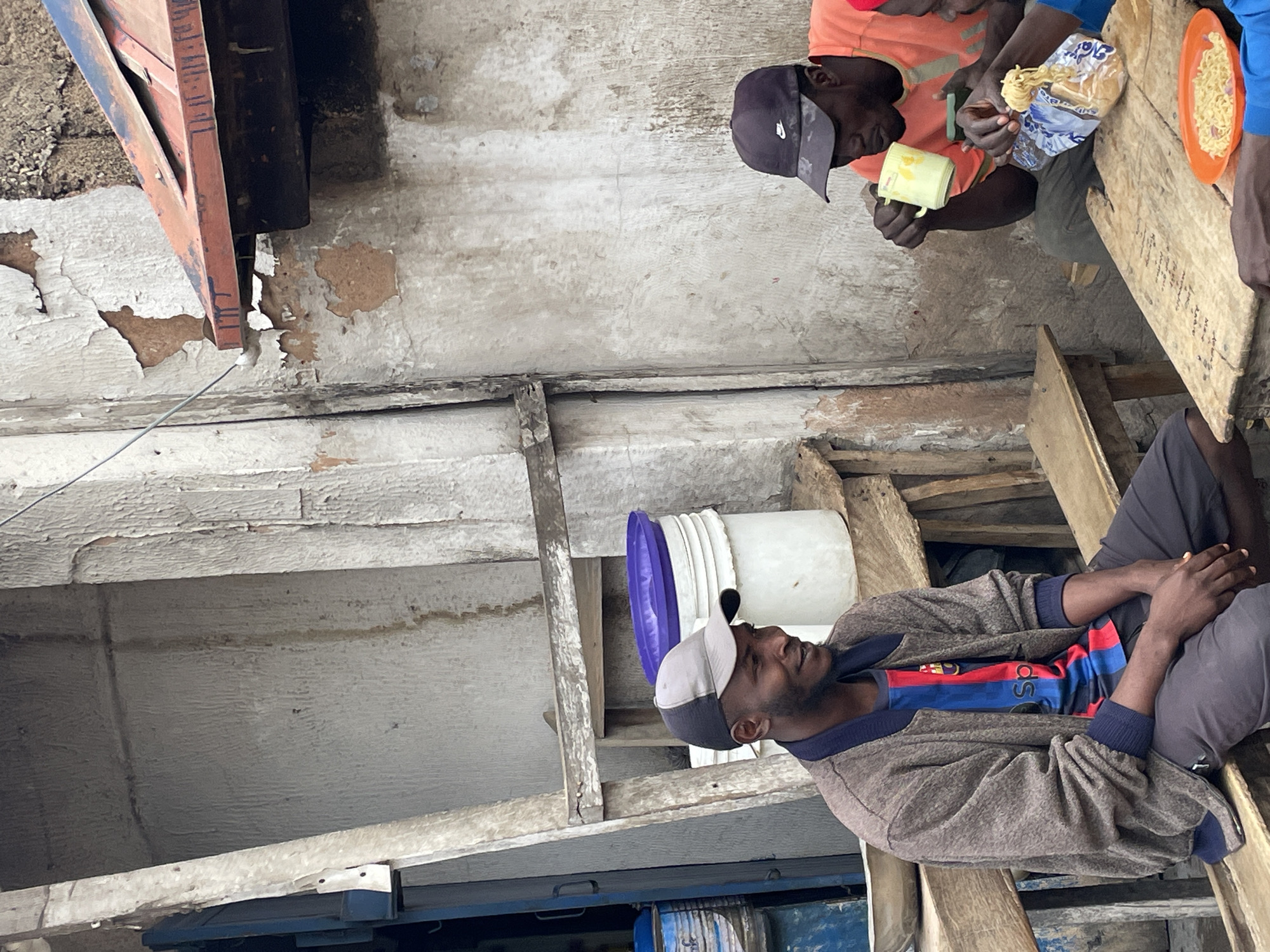 The image shows a scene in Jos, Nigeria. Two men are seated at a wooden table, eating. One man wears an orange shirt and a dark baseball cap and is holding a yellow cup and a package of food. Another man's hand is visible near an orange plate with noodles. A third man sits nearby on wooden planks. He is wearing a grey jacket, a shirt with red and blue stripes, dark pants, and a two-tone baseball cap. A white bucket with a purple lid is behind him. The background includes distressed walls with peeling paint and wooden beams. There are blue containers visible in the lower part of the frame.