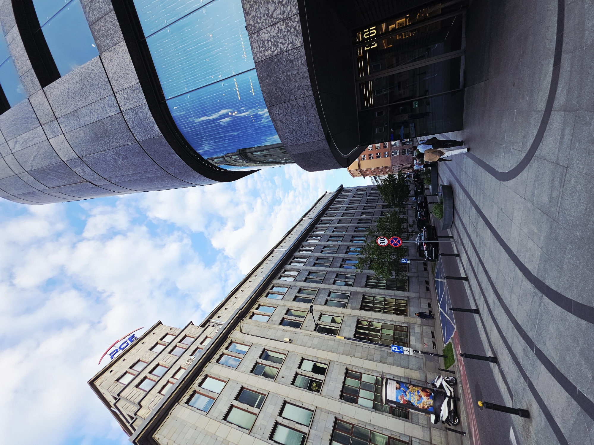 The image shows an exterior scene in Warsaw, Poland, featuring two prominent buildings under a blue sky with scattered clouds. One building has a curved, glass-paneled facade reflecting the sky, with a textured gray stone exterior and visible signage reading "25." The other building is a taller, older structure with many windows and a sign reading "PGE" on its roof. A street runs between the buildings, with visible pedestrian traffic, including people walking. There are street signs and a parked scooter. The foreground features a paved area with discernible patterns.