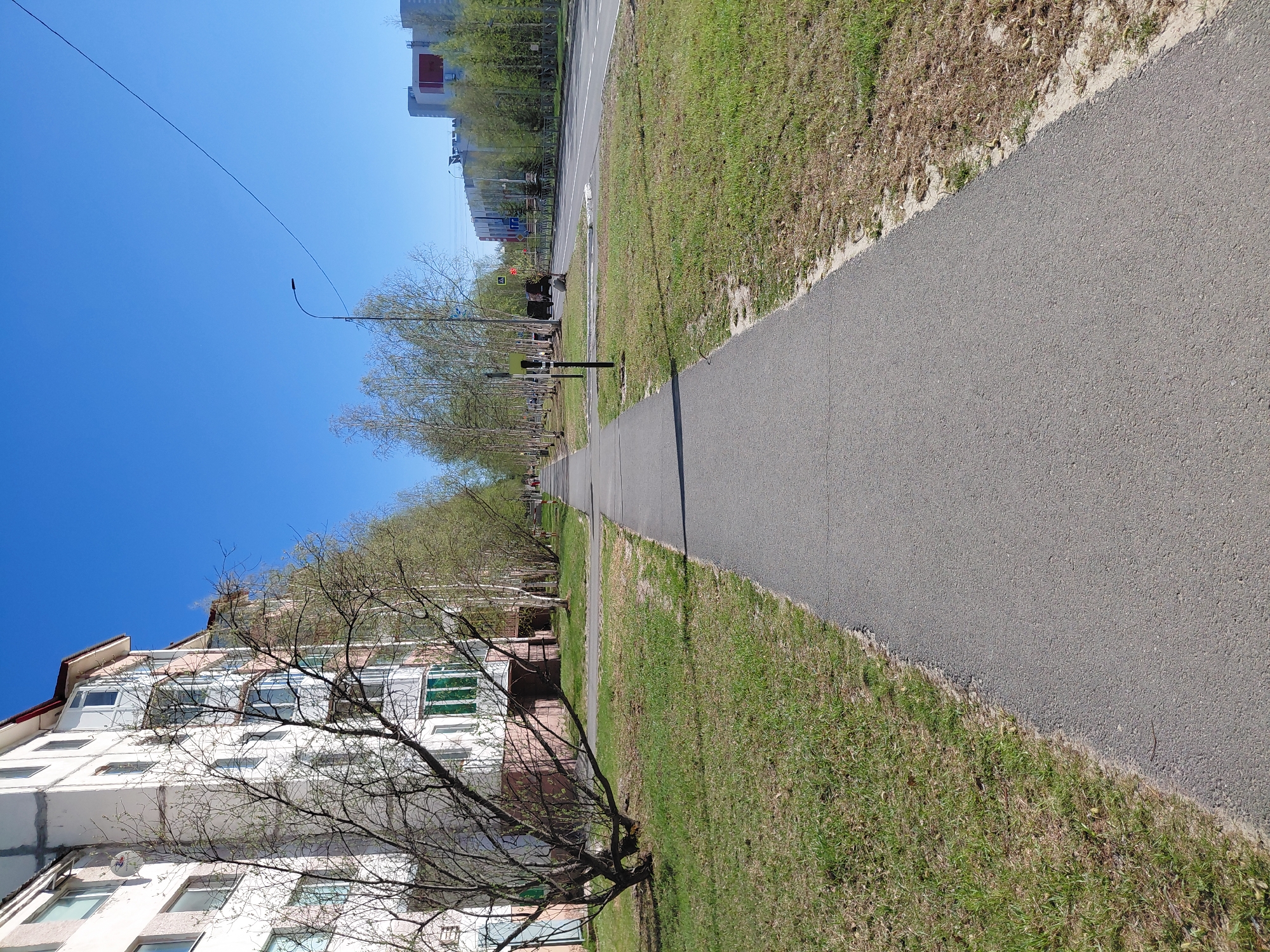 The image, taken in Nadym, Russia, shows an outdoor scene featuring an apartment building, a pedestrian path, and a clear blue sky.
On the left side of the frame, a multi-story residential building with numerous windows and balconies dominates, its white facade visible through the bare branches of several trees positioned in front of it. The trees, mostly deciduous, show signs of early spring with some nascent green leaves, while others remain largely bare.
To the right of the trees, a paved asphalt path or narrow road stretches into the distance, appearing to run parallel to the building. This path is bordered on both sides by grassy strips, featuring a mix of green and dry, brown grass. Further to the right, beyond the path and grass, there appears to be another, wider paved road or thoroughfare, also lined with grass and trees.
In the far distance along the path, several indistinct human figures can be seen, engaged in walking, suggesting routine daily activity. Streetlights and overhead power lines are visible, extending into the clear blue sky.
The scene is set during bright daylight, with a stark, cloudless blue sky overhead and strong sunlight casting distinct shadows. The overall impression is that of a calm, everyday spring day in a residential area. No legible text is visible in the image.