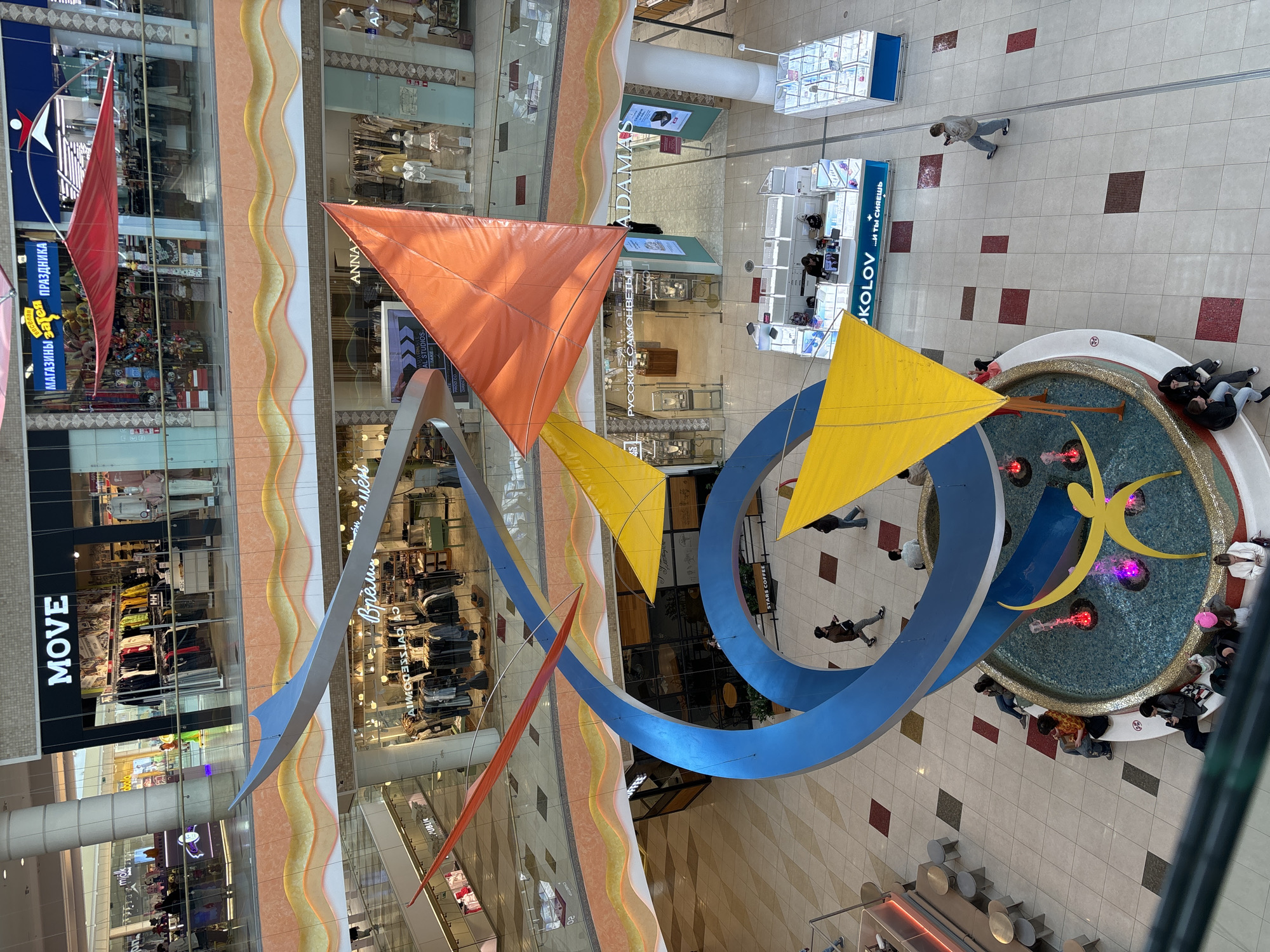 Captured from an elevated viewpoint, the image portrays the interior of a shopping mall located in an unidentified city. The scene is dominated by a hanging art installation featuring orange, yellow, and red triangular "sails" along with curving metal structures, the largest of which is a blue "C" shape. The artwork is suspended above the mall's open area, offering views down to the ground floor and across to the storefronts of different levels.
Storefronts such as "MOVE," "ANNA," and "ADAMAS," visible through the architectural elements, suggest a variety of shops selling clothing and jewelry. Patrons can be seen browsing within the stores, and a person walks through the open area below.
A central fountain surrounded by people is visible on the ground floor, adding a focal point to the scene. The mall's interior design includes a tiled floor and wavy architectural elements on the walls of the different levels.
Overall, the photograph depicts a typical shopping mall environment, complete with shops, art installations, and the flow of customers within the space.