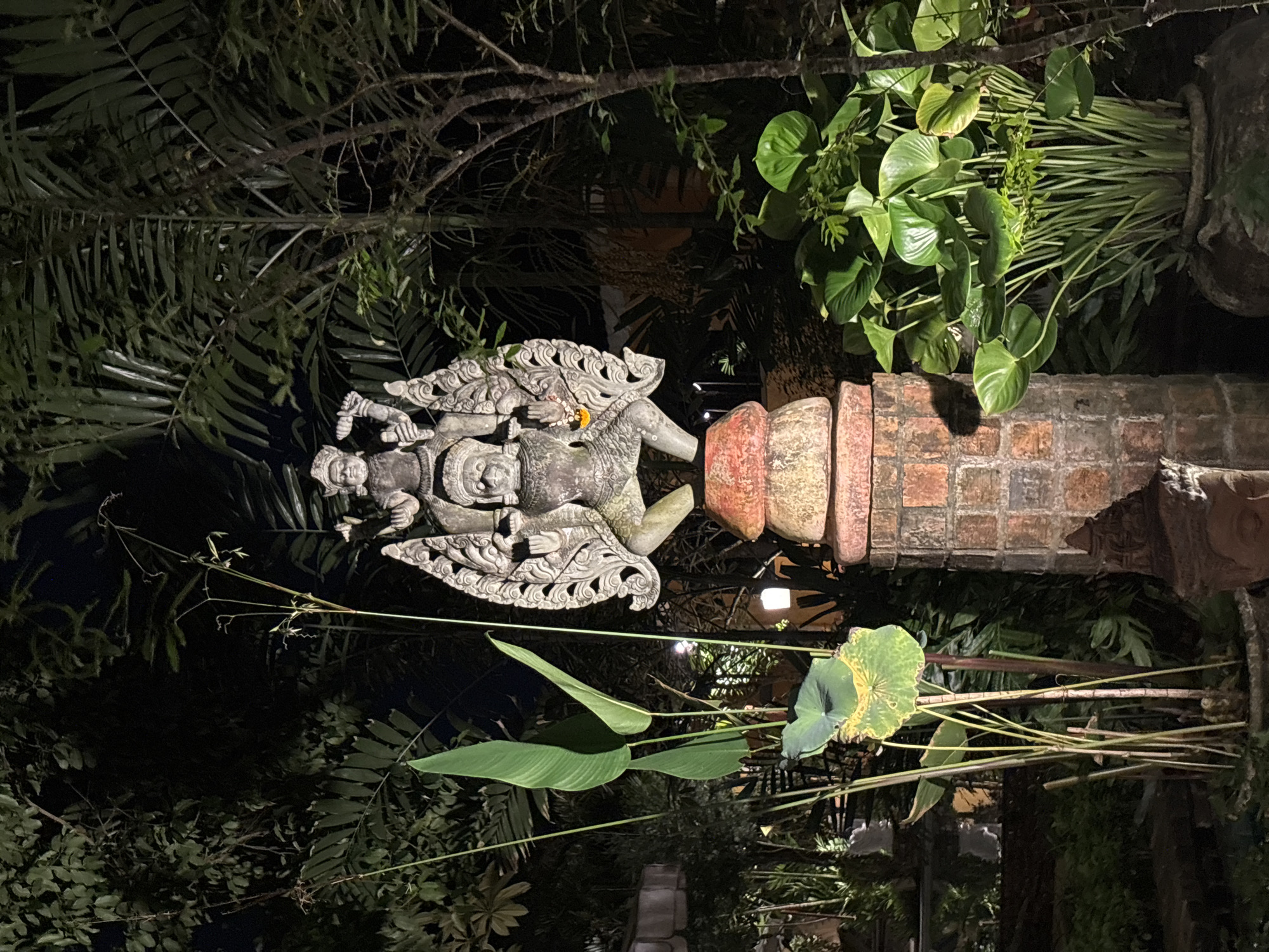 A nighttime shot captures a stone statue of a deity, likely from Southeast Asian mythology, positioned on a brick pedestal. The statue depicts a figure with elaborate headdress and intricate carvings, holding a weapon in its right hand. It is surrounded by lush green foliage and palm trees, illuminated by artificial lights that cast shadows and highlight the textures of the stone and plants. In the background, glimpses of buildings and outdoor lighting suggest an urban or resort setting, possibly within the city of Chalong, Thailand. The overall ambiance is serene and somewhat mystical, with the darkness of the night contrasting with the illuminated subjects.