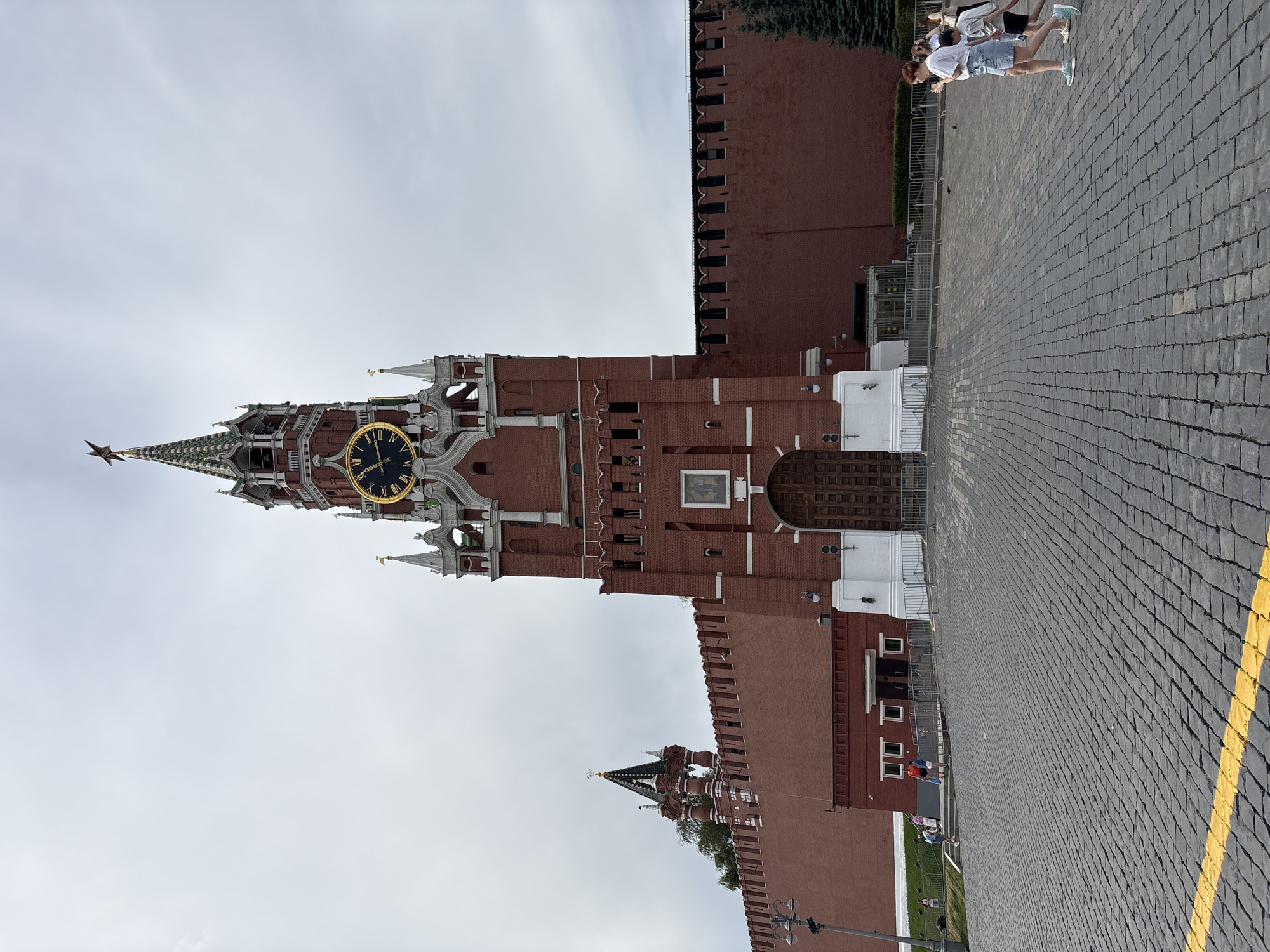 Here is a description of the image:
The image captures the Spasskaya Tower, a prominent landmark of the Moscow Kremlin in Russia. The tower, constructed of red brick and white stone, features a large clock face and a red star atop its spire. To the right of the tower, a section of the Kremlin wall is visible, constructed of similar red brick. The foreground is dominated by a paved area with a yellow line along its edge. Several individuals are visible walking in the upper right corner of the image. The sky is overcast.