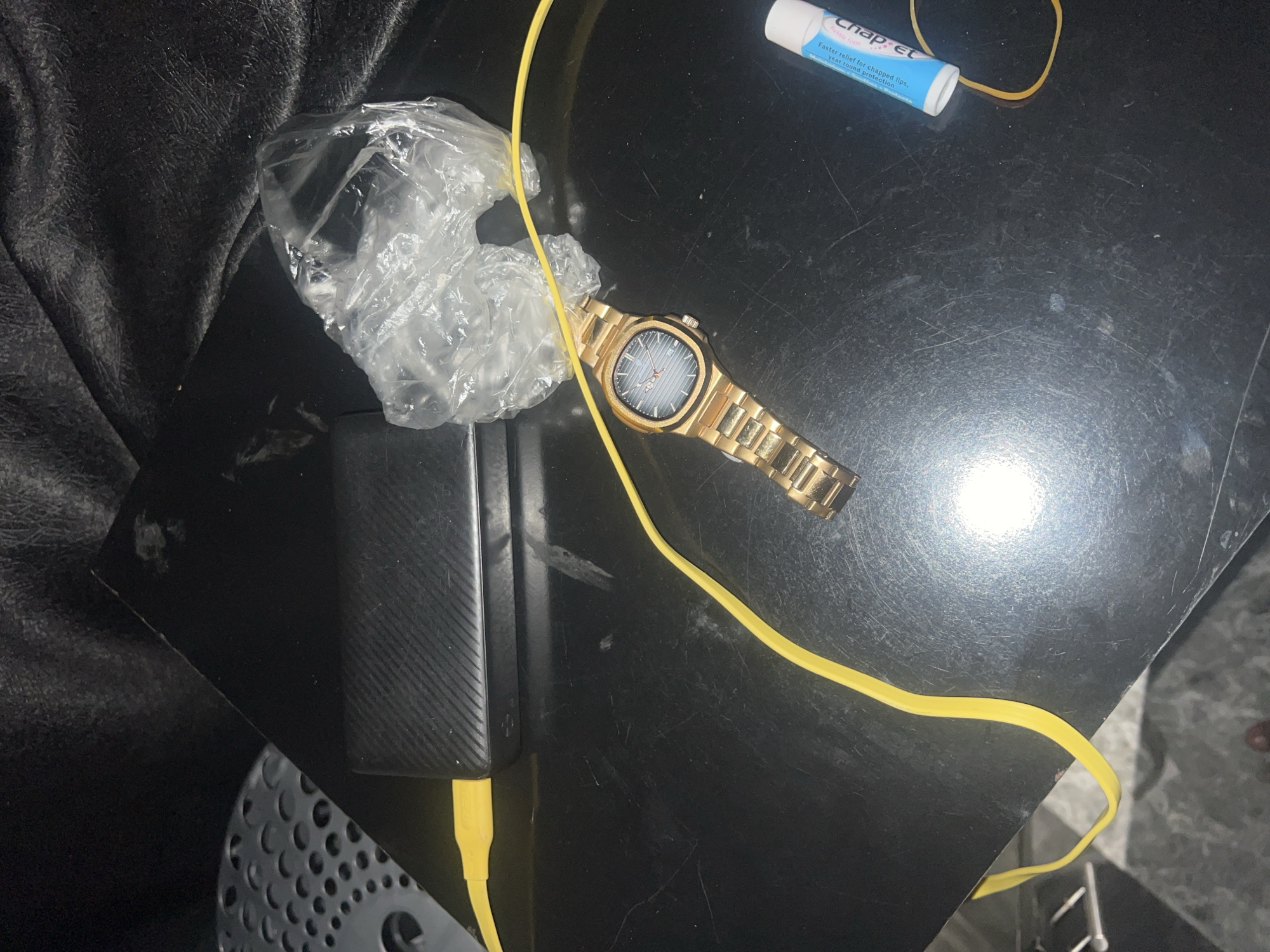 The scene depicts various personal items arranged on a dark, scratched, and reflective flat surface. A black power bank, featuring a textured, diagonal line pattern reminiscent of carbon fiber, is connected on its left side to a bright yellow USB cable. This cable extends across the surface, curving past a crumpled, transparent plastic bag situated in the upper-right portion of the frame. A gold-colored wristwatch, equipped with a metal link band and a blue dial marked with horizontal lines, is positioned centrally. In the lower right corner, a white and blue tube of "chap-ex" lip balm is visible, alongside a light brown rubber band. The upper-left background displays dark, textured material, potentially leather or fabric, with a grey, perforated object partially visible on the far left. No individuals or specific actions are depicted. The image contains no visual information or identifiable landmarks to indicate the requested location of Jauro Yinu, Nigeria.