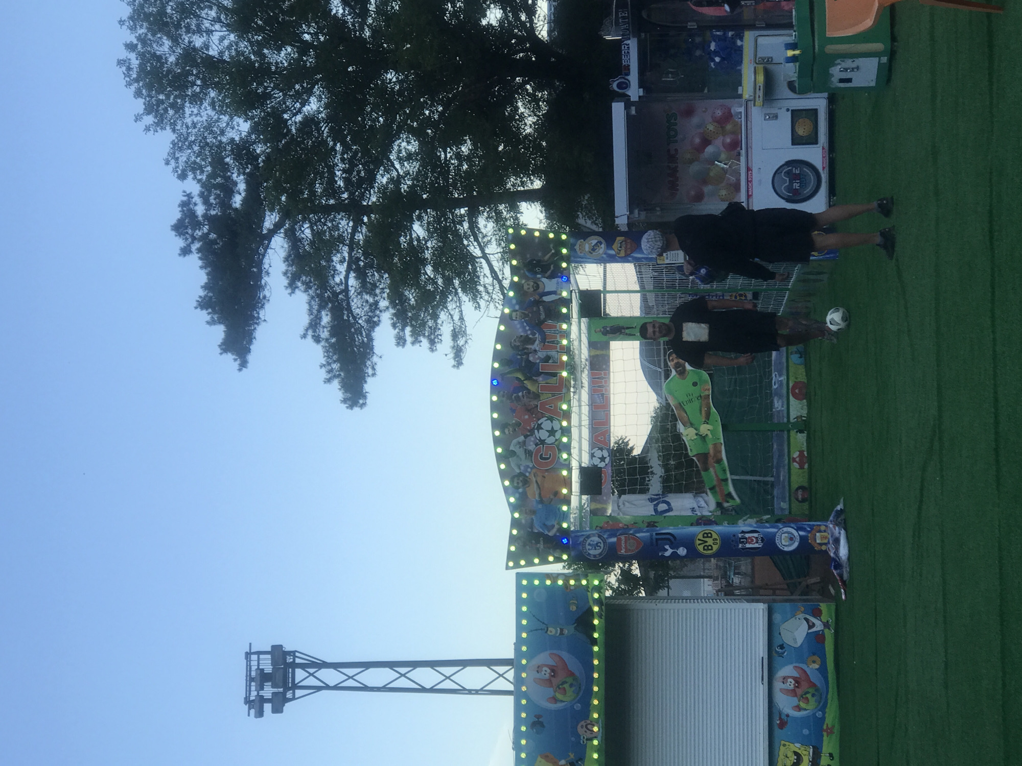 This photograph captures an outdoor scene at a fairground, likely in Varna, Bulgaria. The image is oriented vertically, with the sky and trees dominating the upper portion and a green turf-like surface covering the bottom.
The main subject is a carnival game booth designed with a soccer theme. The booth features colorful graphics of soccer players and the word "GOALL!!!" prominently displayed. Adjacent to the game booth, there are two individuals, one standing and another slightly behind him, both appearing to be dressed in dark clothing. A soccer ball is visible on the green surface in front of them.
Behind the soccer game, there is a "MAGIC TOYS" vending machine with rows of colorful balls. To the left of the booth, a tall metal structure with lights, possibly part of a larger ride or attraction, is visible against the bright blue sky. The time of day appears to be daytime, with ample natural light. The overall atmosphere seems casual and recreational, typical of a fair or amusement park setting. Visible text includes "GOALL!!!", "MAGIC TOYS", "MAGIC TOTS", "BVB", "Arsenal", and "BEARDATE".