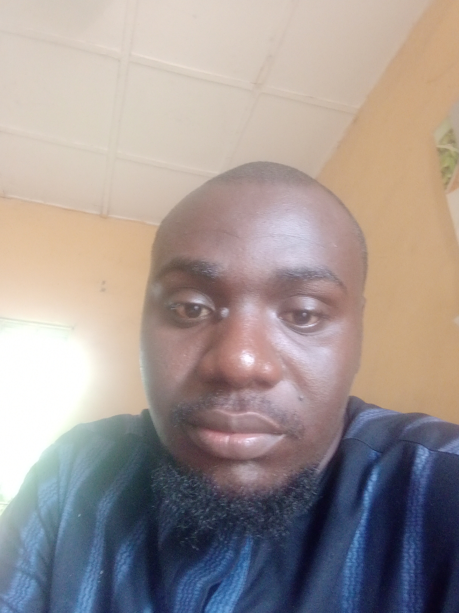 This is a medium-shot, waist-up selfie of a dark-skinned man. He is looking directly at the camera, with a neutral expression. The man is bald, and has a beard and mustache. He is wearing a dark blue and black patterned shirt with a high neckline.
The background is an indoor setting, with a yellow wall and a white ceiling that appears to be made of panels. There is a window to the left of the frame, which is emitting a bright light, suggesting it is daytime. The overall lighting in the image is somewhat uneven, with some areas brighter than others. No text is visible in the image. The context of Jalingo, Nigeria, suggests a potentially warm climate, which may influence the clothing choice.
