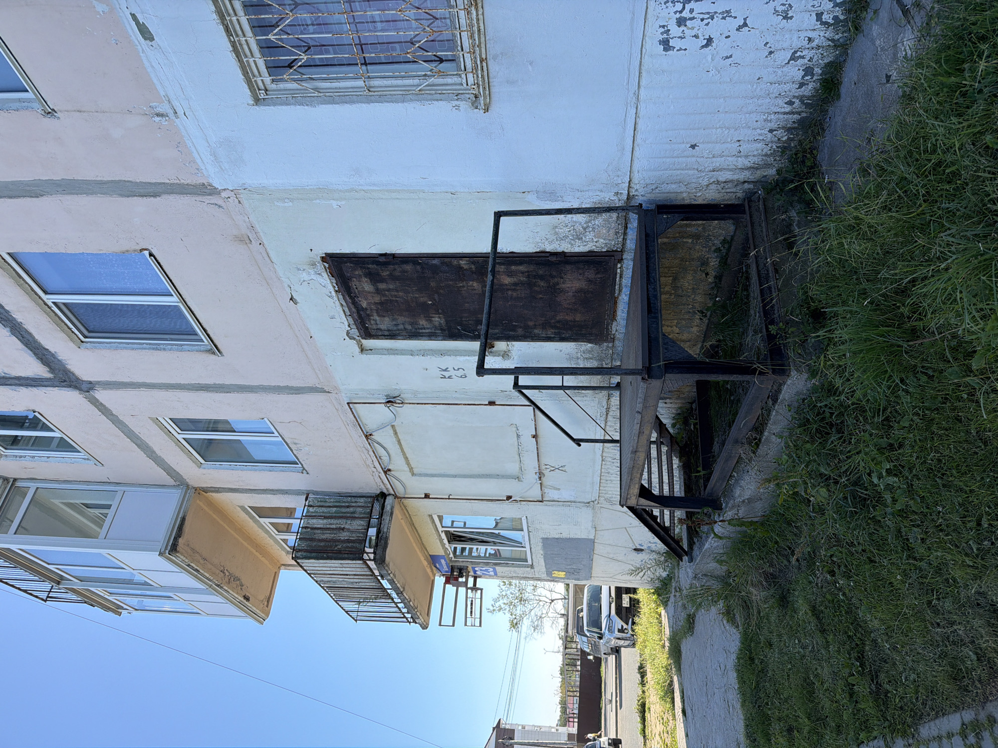 An exterior view of a light-colored apartment building is captured under a clear blue sky. The building has multiple windows, some with white frames and others with protective metal bars. A metal balcony railing, showing signs of rust, is visible on an upper floor, as is a glass-enclosed balcony. A dark, weathered metal door is situated at ground level, accessed by a set of metal stairs and a small platform. The building's facade exhibits peeling paint and some visible concrete textures. The foreground features a patch of unkempt grass bordering a concrete walkway. In the background, a street scene unfolds with vehicles, including a white SUV with a license plate that appears to be from Russia, and other buildings. The location is identified as Okha, Russia, specifically the building at 23 Berezovaya Street. The scene conveys a sense of urban, possibly aging, infrastructure.