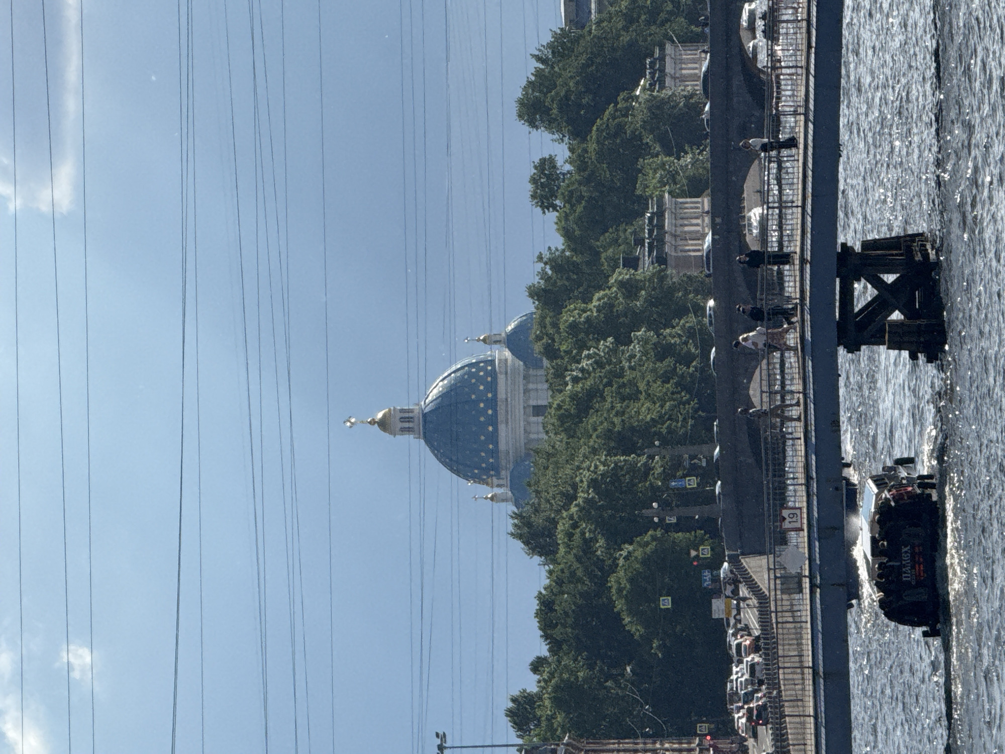 The image captures a view of the Trinity Cathedral in Saint Petersburg, Russia, as seen from across the Neva River. The cathedral, with its distinctive blue dome adorned with gold stars and topped by a golden cross, is the central focus of the background. It is surrounded by a dense canopy of lush green trees. In the foreground, a pedestrian bridge spans the river, with several people walking along it. Below the bridge, an amphibious vehicle, possibly a tour boat, navigates the choppy water. The vehicle has "ПАЛЕХ" written in red Cyrillic letters on its rear. In the far background, more of the city's architecture is visible, along with traffic on a road adjacent to the river. Overhead, a network of power lines crisscrosses the sky, interrupting the view of the blue sky and scattered clouds. The overall scene depicts a sunny day in the city, with a blend of historical architecture, natural elements, and urban activity.