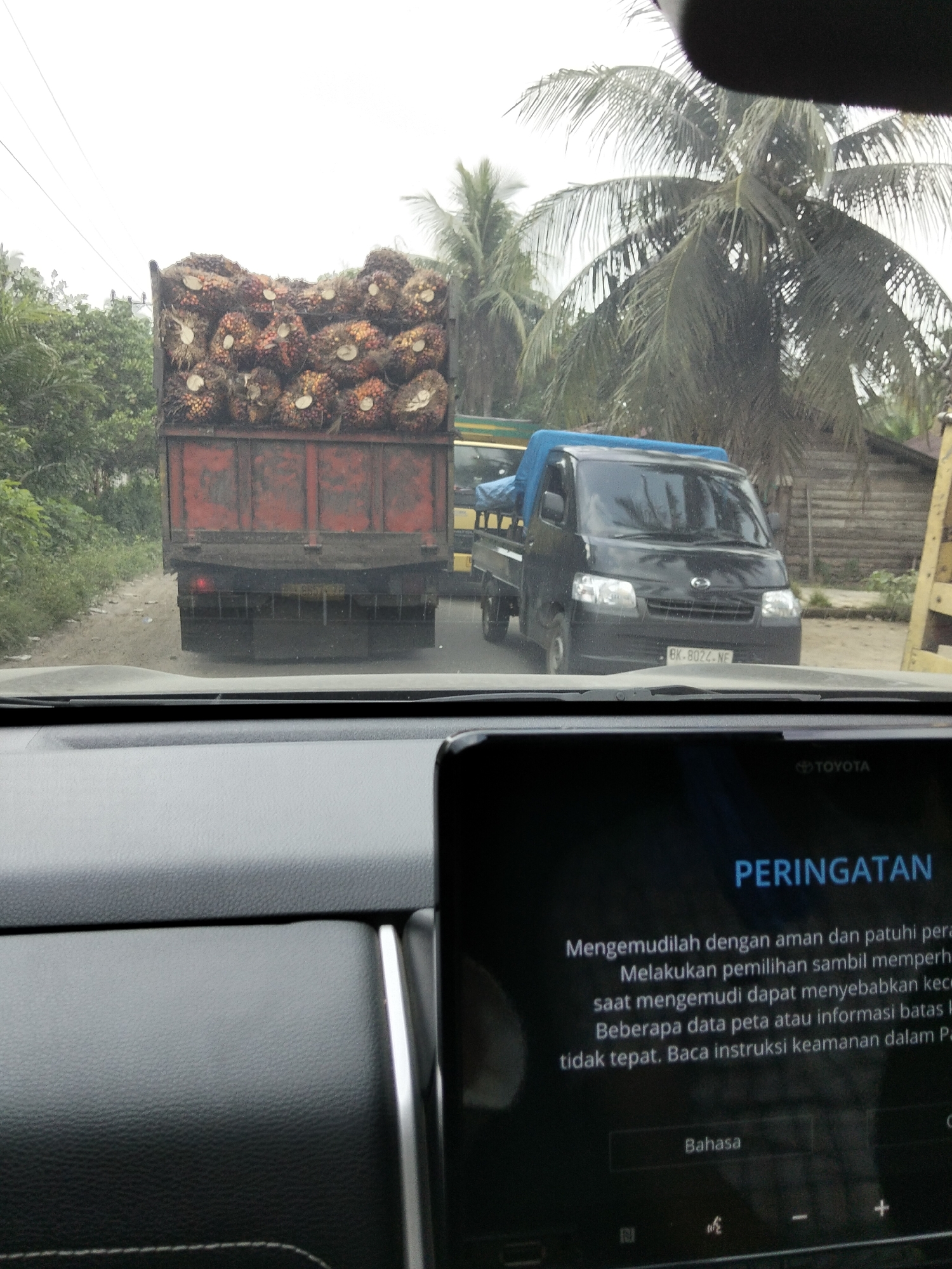 This is a photo taken from inside a vehicle in Awegeutahpaya, Indonesia. The vehicle is on a dirt road with some vegetation on the left side. In front of the vehicle, there is a red truck loaded with palm oil fruit. To the right of the red truck, there is a black pickup truck with a blue tarp covering its bed. The pickup truck has a license plate that reads "BK 8022 NF". In the background, there are palm trees and wooden structures. The foreground of the image shows the dashboard of the vehicle the photo was taken from, with a car infotainment screen displaying a warning message in Indonesian. The screen shows the Toyota logo at the top.