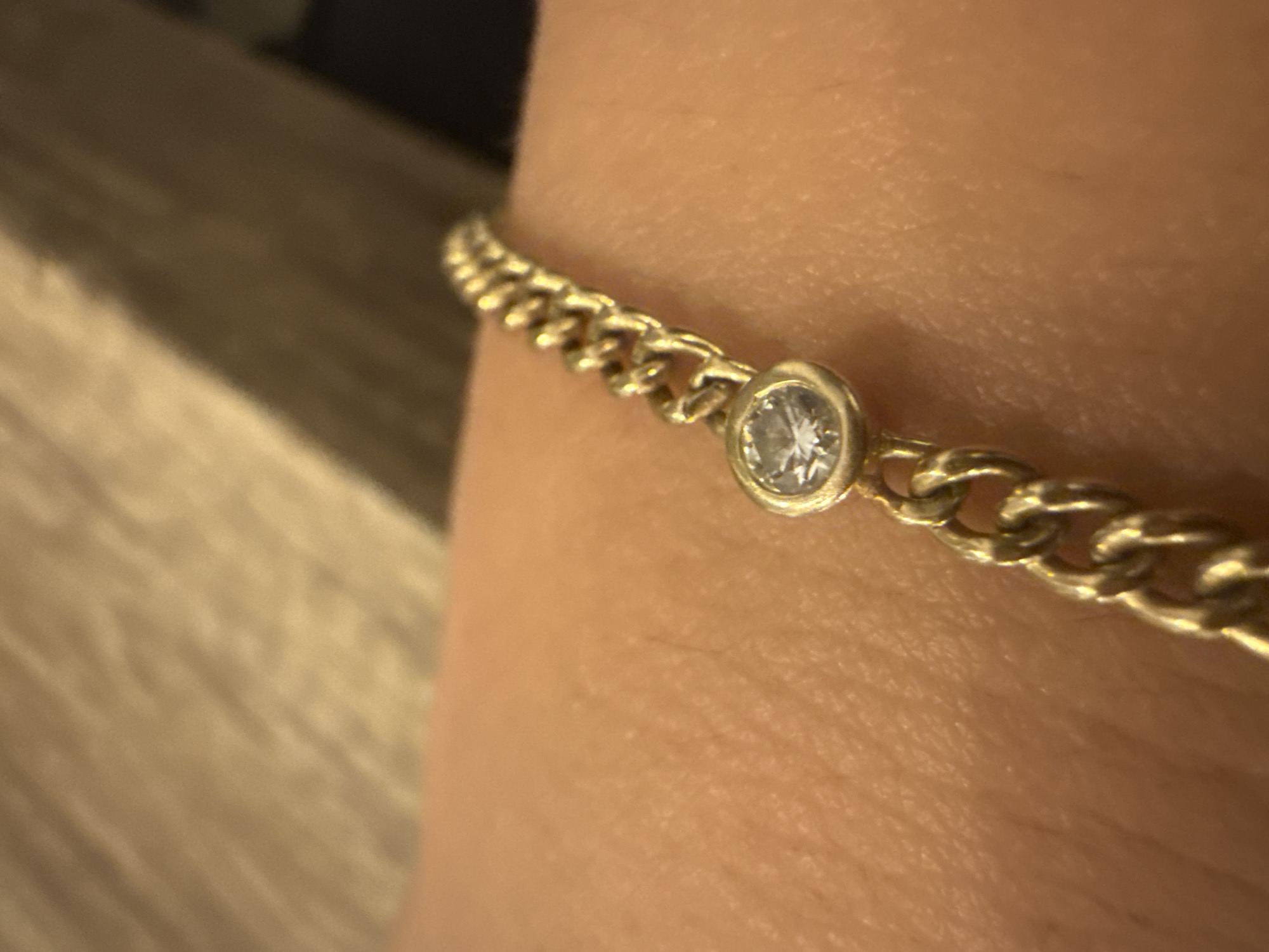 A gold curb chain bracelet with a single round-cut cubic zirconia gemstone is resting on a person's skin. The bracelet is positioned diagonally across the frame. The background is blurred, appearing to be a textured surface. The location is not specified in the image or provided information.