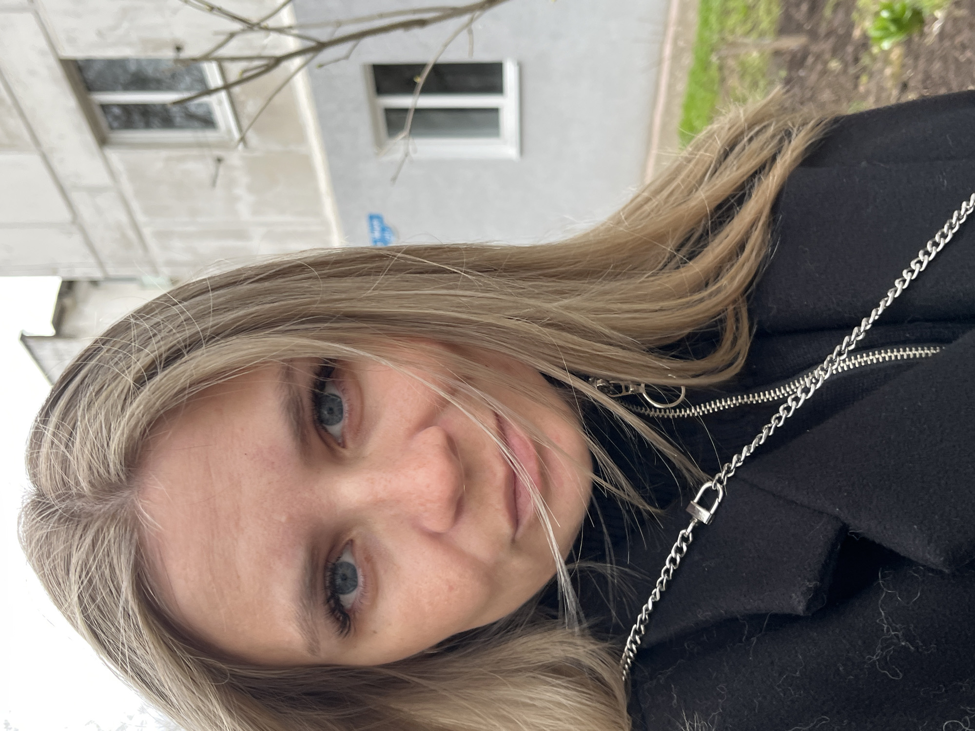 An angled, close-up self-portrait features a young woman outdoors in Chaikovsky, Russia. She has long, light blonde hair, fair skin, and blue eyes, and looks directly at the camera with a subtle, soft smile. She is wearing a black coat or jacket, detailed with a visible silver zipper and a decorative silver chain that includes a small, padlock-shaped charm. The background is softly blurred but indicates an urban setting, showing a light-colored building with a rectangular window and several bare tree branches, suggesting a late winter or early spring season. The overall lighting is soft, likely due to overcast conditions. There is no visible text in the image.