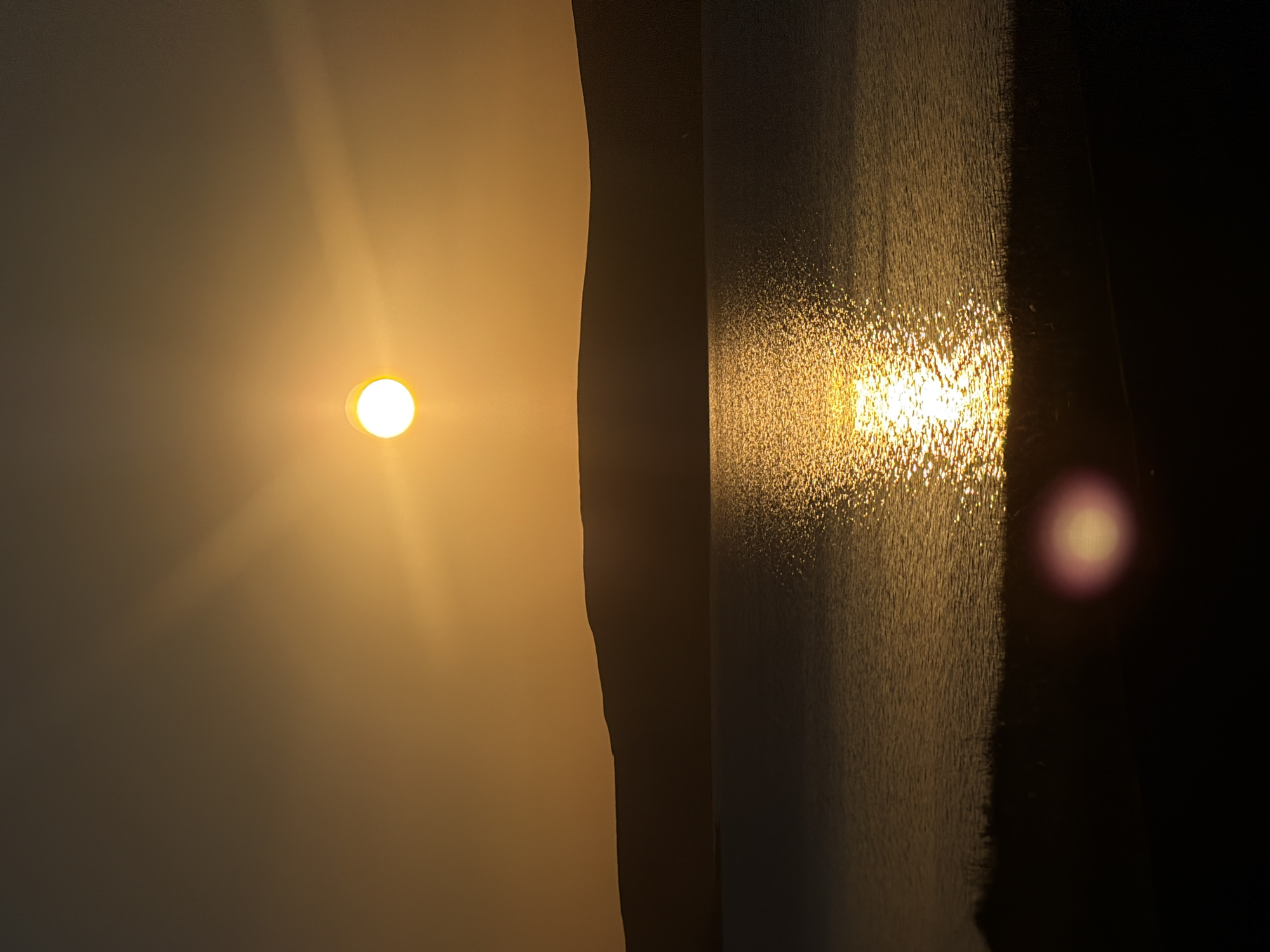 The image captures a serene sunset over a body of water. The sun, a bright orb, is positioned in the upper portion of the frame, casting a warm, golden glow across the sky and reflecting brilliantly on the water's surface. A silhouetted hillside forms the horizon, creating a stark contrast against the vibrant sky. The foreground is dark, with the silhouette of vegetation visible along the water's edge. A lens flare, appearing as a soft, circular light, is present in the lower portion of the image. The scene appears to be an outdoor natural landscape.