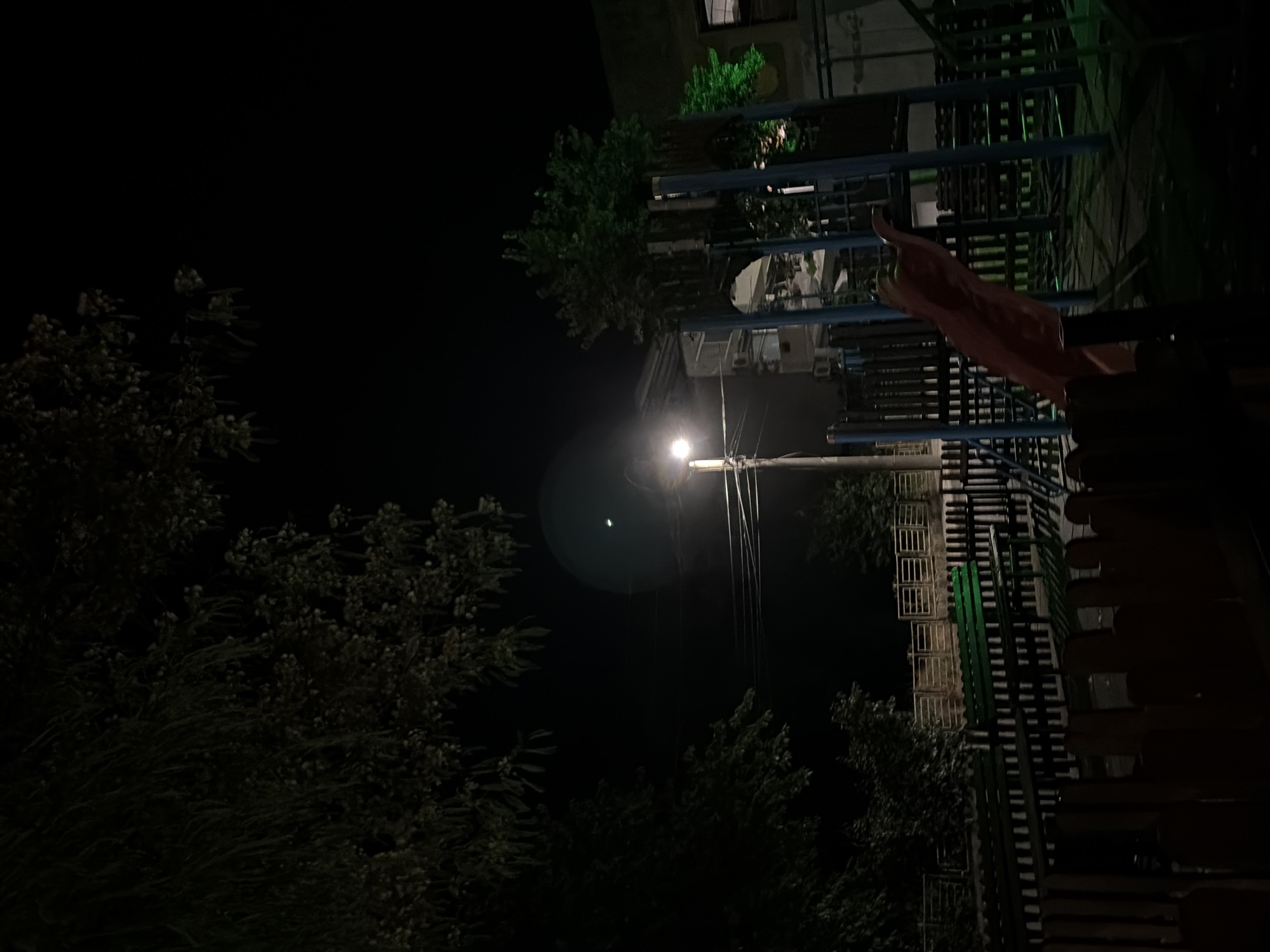 An outdoor playground is depicted at night in Obzor, Bulgaria. The scene is illuminated by a streetlamp and some green lights from a building. In the foreground, a dark wooden fence is visible. Behind the fence are two green park benches and a low fence. To the right, a blue and orange playground structure with a red slide is prominent. In the background, trees and a building with green lights are present. Power lines are strung between poles, with one pole supporting the streetlamp. The sky is dark, indicating nighttime.