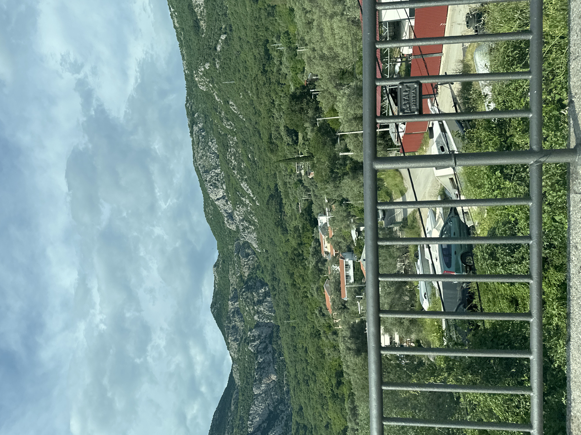 A mountain covered in vegetation is viewed through a metal railing. A small village with houses is located on the mountain's slope. The sky is cloudy. The location is Kaluđerac, Montenegro. Parked cars and boats are visible behind the railing.