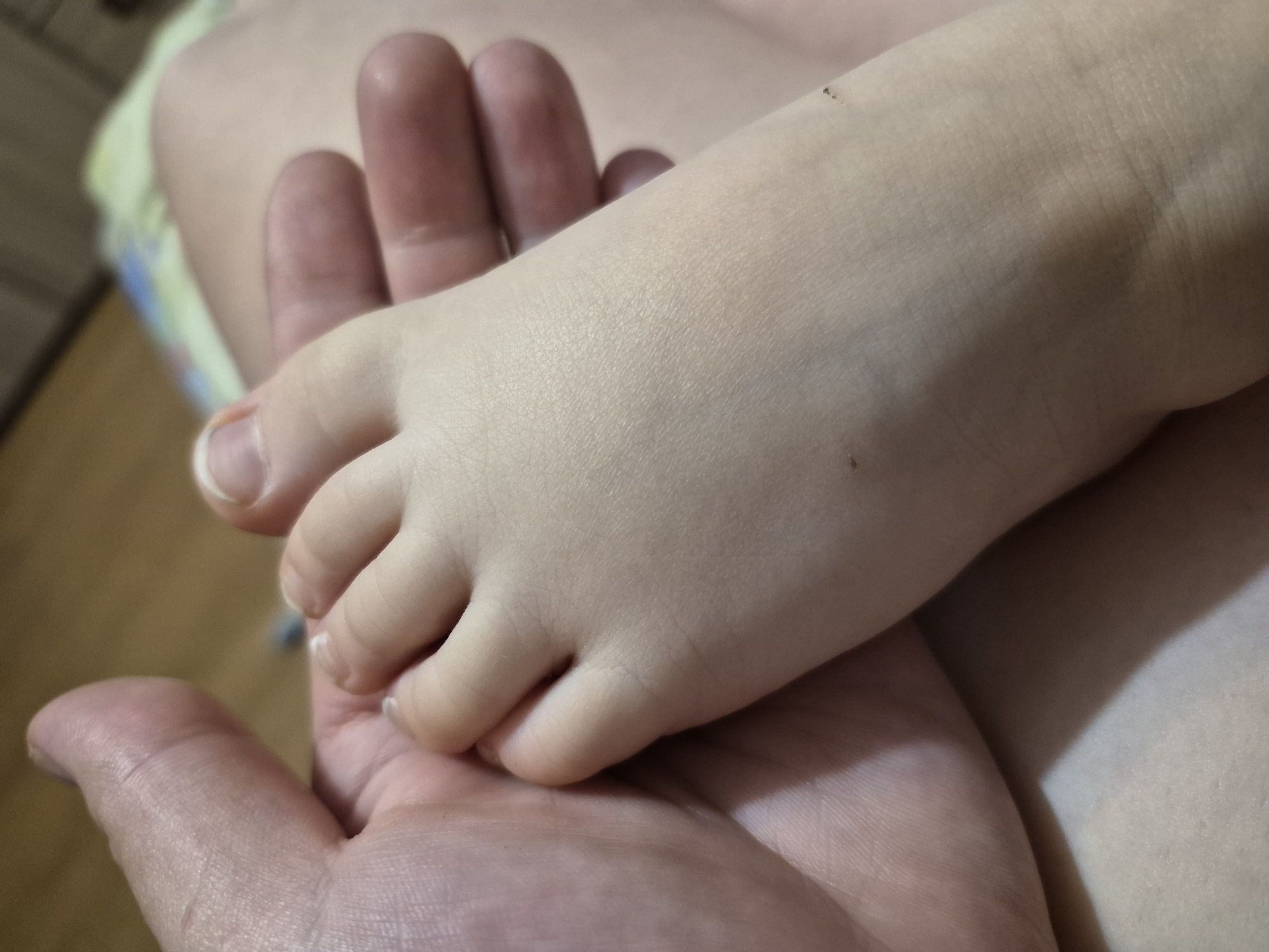 A close-up, intimate photograph captures a tender moment between an adult and a child in what appears to be an indoor setting in Chaikovsky, Russia. The primary subjects are a large, open adult hand and a small, delicate child's foot resting within it.
The adult's hand is seen from the palm-up perspective, showing visible skin texture and creases, suggesting a mature hand. The fingers are slightly curled, providing a gentle cradle for the child's foot. The child's foot is significantly smaller, with smooth, fair skin and soft, rounded toes. The toenails are visible and appear neatly trimmed. A couple of tiny, dark specks are noticeable on the top of the child's foot.
The lighting is soft and warm, casting gentle shadows that highlight the contours of the hands and foot. The background is out of focus, showing blurred shapes and soft, light colors, possibly indicating clothing or bedding, but no distinct objects or location cues are identifiable beyond the general indoor environment.
The image conveys a strong sense of care, protection, and connection between the two individuals. The act of the adult holding the child's foot suggests a moment of comfort, affection, or quiet intimacy. No visible text is present in the image.