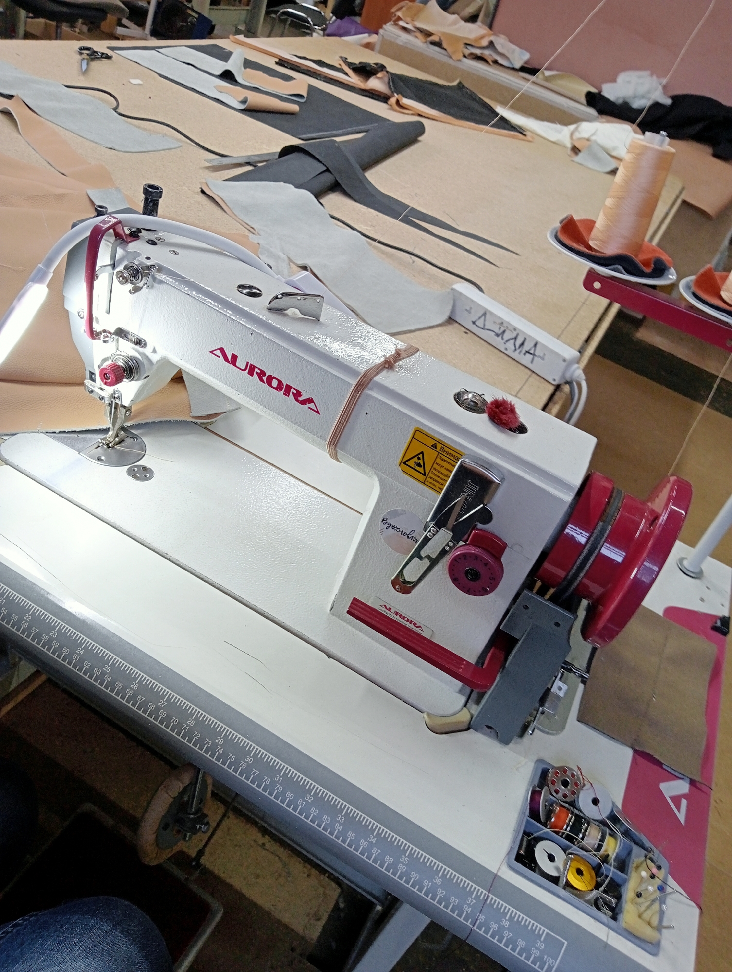 This is a medium-shot, eye-level, interior shot of a sewing machine in operation at a workshop. The sewing machine is white with red accents and features the brand name "AURORA" in red letters. A light is attached to the sewing machine's arm. Various pieces of leather and fabric are laid out on a light-colored wooden table in the background, suggesting a workshop environment. There is a spool of orange thread on the right side of the image, and a container of smaller thread spools, bobbins, and needles is on the table in the foreground. A ruler with centimeter markings is attached to the edge of the table. The scene appears to be in Saint Petersburg, Russia, possibly a small business or home studio.