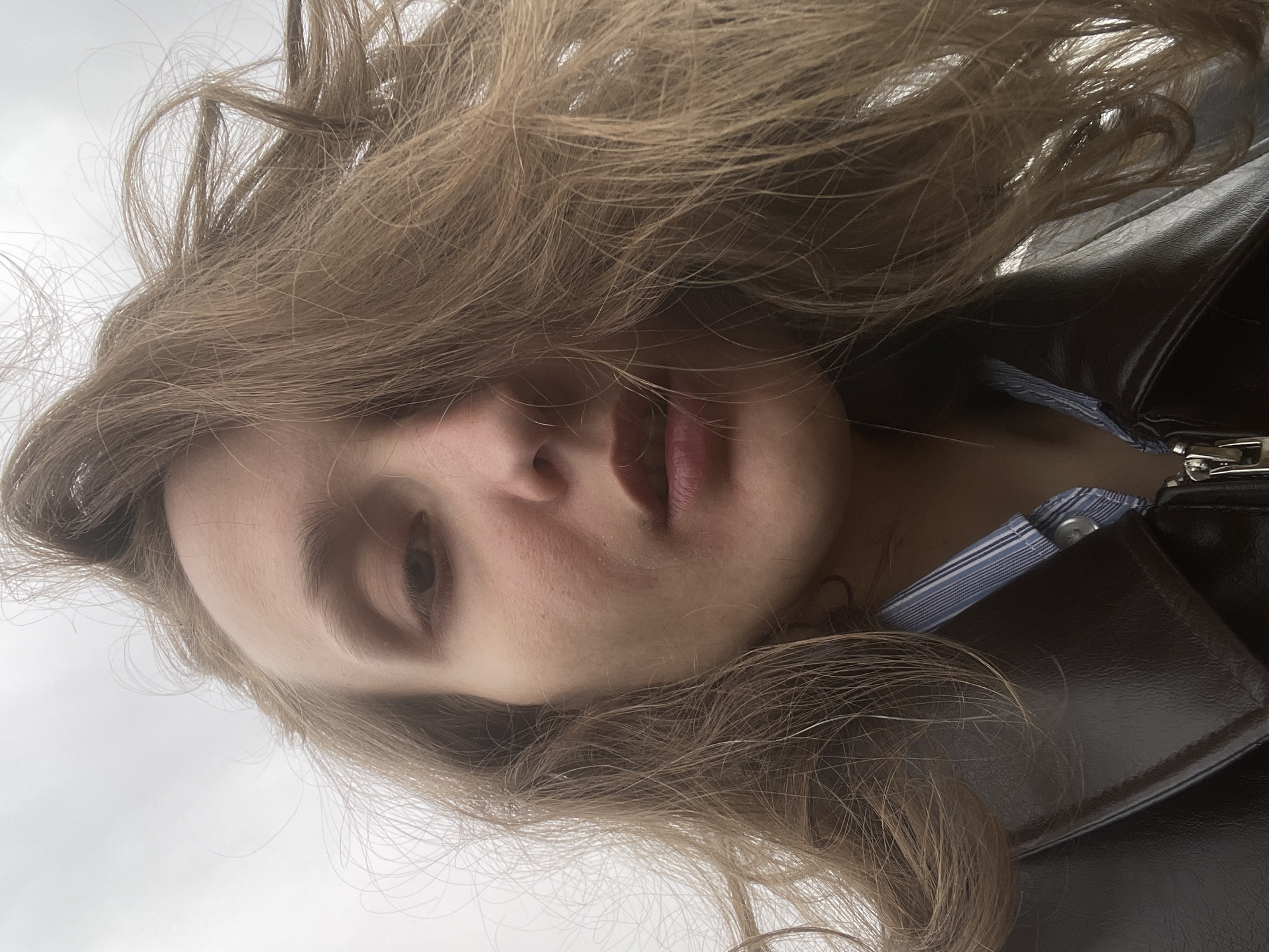 A close-up, slightly rotated view of a person's face with long, brown hair partially obscuring the forehead and face. The person has light skin and one eye is visible. The person is wearing a brown leather jacket over a blue and white striped shirt. The background is a cloudy sky. The location is unknown, so I cannot confirm its presence in Подшивалово, Russia.