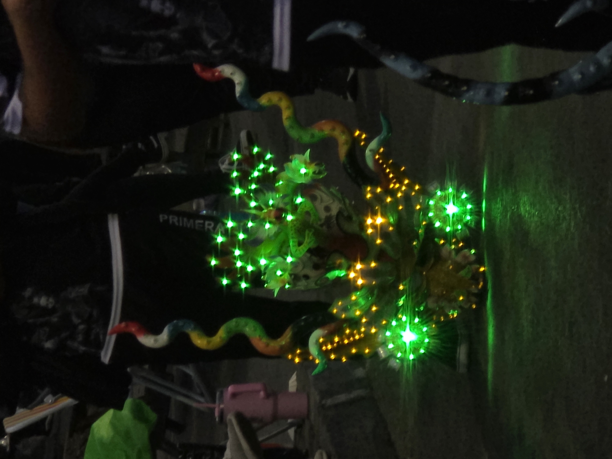The image depicts a nighttime scene outdoors, likely on a street or public area, in Iquique, Chile. The central subject is a brightly lit, ornate float or display, possibly for a parade or festival. It features a multi-colored, serpentine structure, reminiscent of a dragon or mythical creature, adorned with numerous green and yellow LED lights. The lights create a vibrant glow and illuminate the immediate surroundings. The float is partially obscured by people in the foreground and background, whose faces are not clearly visible due to the low light conditions and their positions. One individual on the right is silhouetted against the scene. The environment appears to be paved, with a darker ground surface. The overall impression is one of a festive or celebratory event at night.