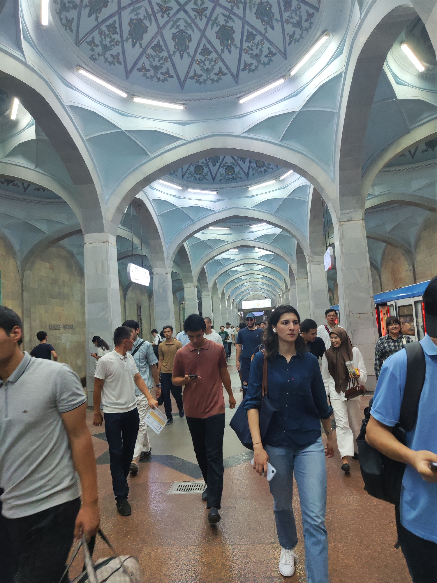 This is a low-angle, eye-level medium shot of a busy subway station with people walking in various directions. The station features a grand architectural style with high, arched ceilings adorned with intricate patterns and lighting. The walls appear to be made of stone or tile, and the floor is a speckled reddish-brown.
In the foreground, a man in a grey polo shirt and black pants is walking from left to right, carrying a duffel bag. To his right, a man in a white polo shirt and black pants is walking towards the viewer. A woman in a dark blue shirt and light blue jeans, holding a smartphone and a dark blue bag, is walking towards the right, slightly ahead of the man in the white polo shirt. Behind her, a woman in a light-colored outfit and a brown hijab is walking to the right, holding a cup and a handbag. A man in a brown polo shirt is walking towards the viewer on the right side of the frame, looking at his phone.
Further in the background, more people are visible walking through the station. A blue subway train is partially visible on the right, with its doors open. The overall atmosphere is one of movement and transit within an ornate, public space. The style of the architecture, particularly the vaulted ceilings and decorative elements, suggests it is a station of the Tashkent Metro, likely one of its more ornate stations.