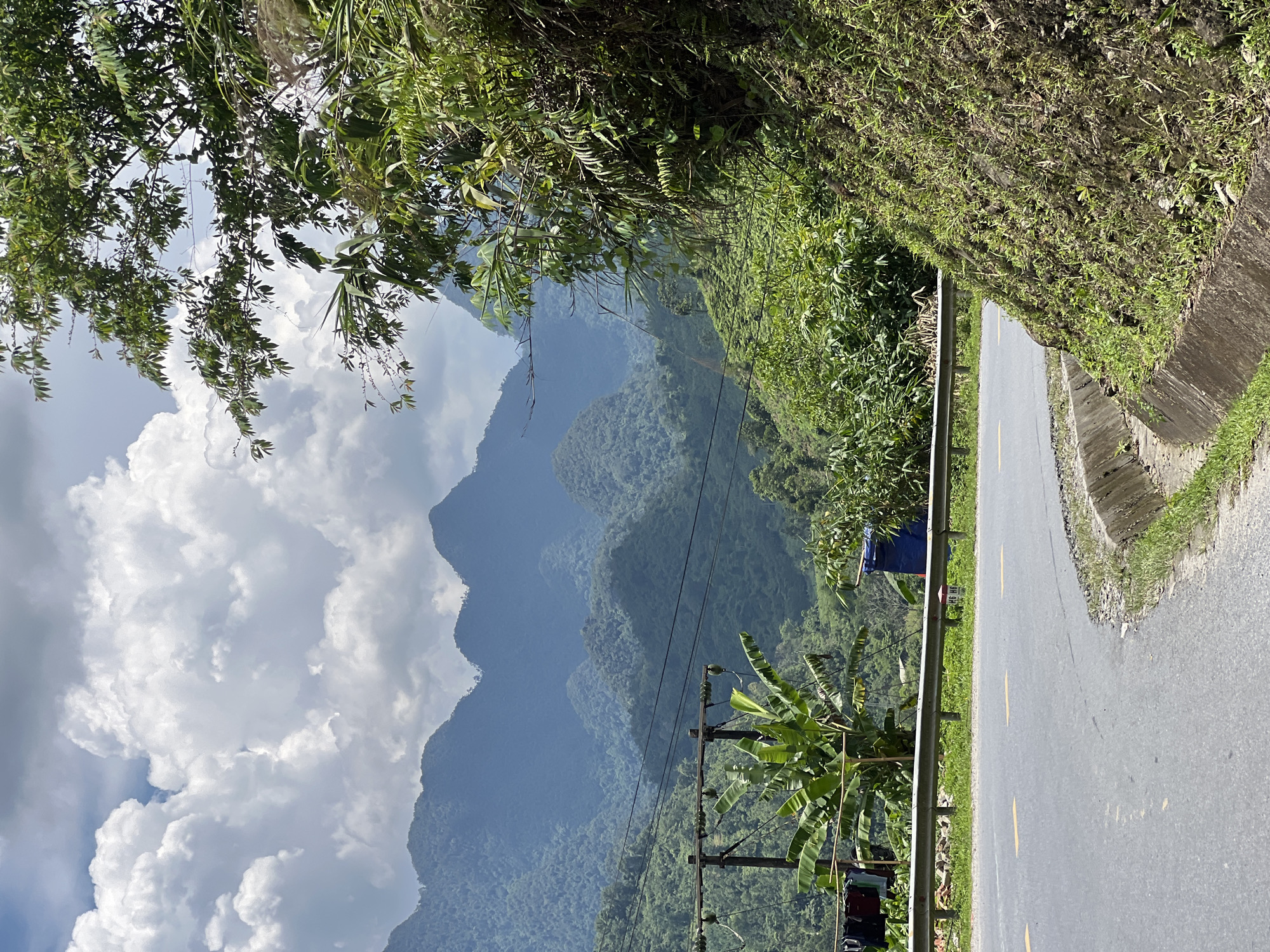 The image depicts a rural mountainous landscape in Minh Tân Commune, Vietnam. A paved road curves through the scene, bordered by a metal guardrail and a vegetated embankment. In the foreground, laundry hangs on a line attached to a utility pole, with a banana tree nearby. Power lines stretch across the middle ground, with a blue tarp visible further down the road. The background is dominated by lush green mountains, layered and receding into a hazy blue distance, under a sky filled with dramatic white clouds. The overall impression is one of natural beauty and a simple, functional roadside environment.