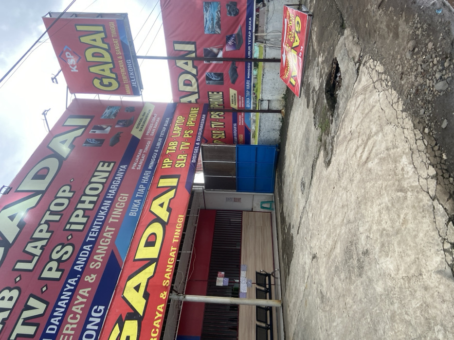 The image shows a pawn shop in Baleendah, Indonesia, located at latitude -7.021 and longitude 107.657. The shop's facade is covered in red and white signage advertising pawn services for electronics, including laptops, TVs, PlayStations, and iPhones. The shop is open, with a blue gate and an open door, and there are four black metal benches lining the wall in front of the shop. The concrete ground in front of the shop is cracked and uneven, with a small hole near the bottom right corner of the image. A blue metal gate is partially open in the background, and an upside-down sign for the pawn shop is lying on the ground. The sky is overcast and gray. The image was taken on a weekday.