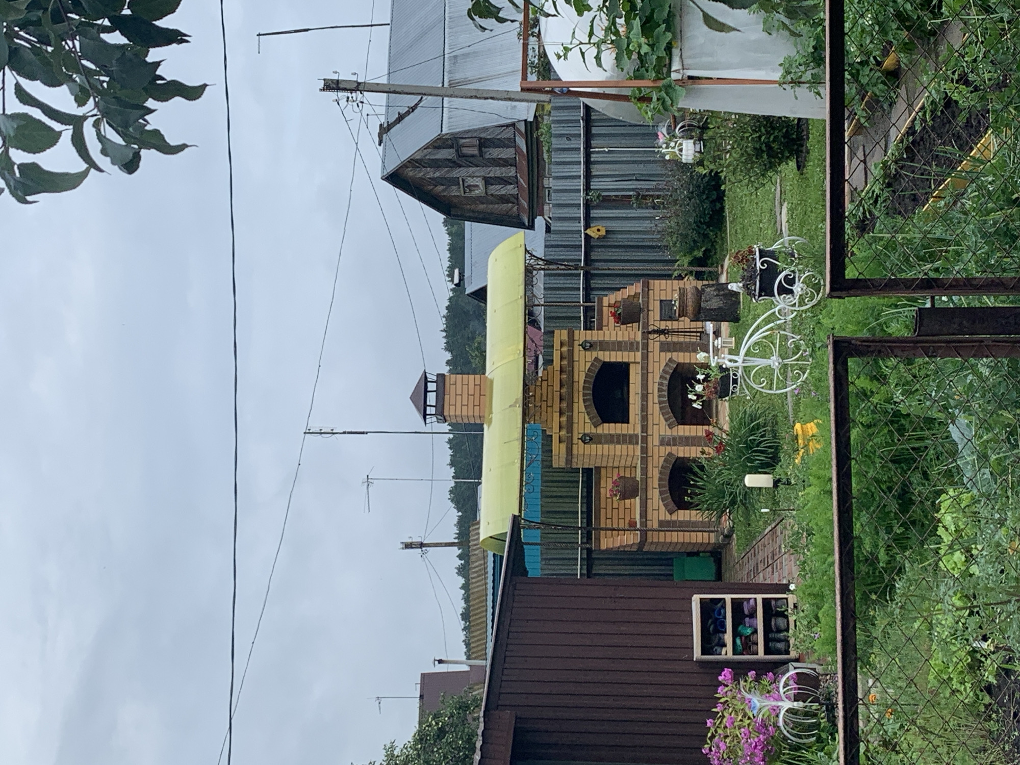 The image depicts a backyard scene, likely in Dubnik, Russia, on an overcast day. In the center, a brick barbecue oven with a yellow canopy is prominent. To its left is a dark brown wooden shed. A white metal decorative bicycle with a planter is positioned in front of the barbecue. Various plants and flowers, including pink and purple blooms, are visible throughout the yard. A metal fence runs across the foreground, separating the viewer from the garden. In the background, several houses with corrugated metal roofs and a greenhouse are visible, along with utility poles and electrical wires. The ground is covered in green grass and a brick pathway.