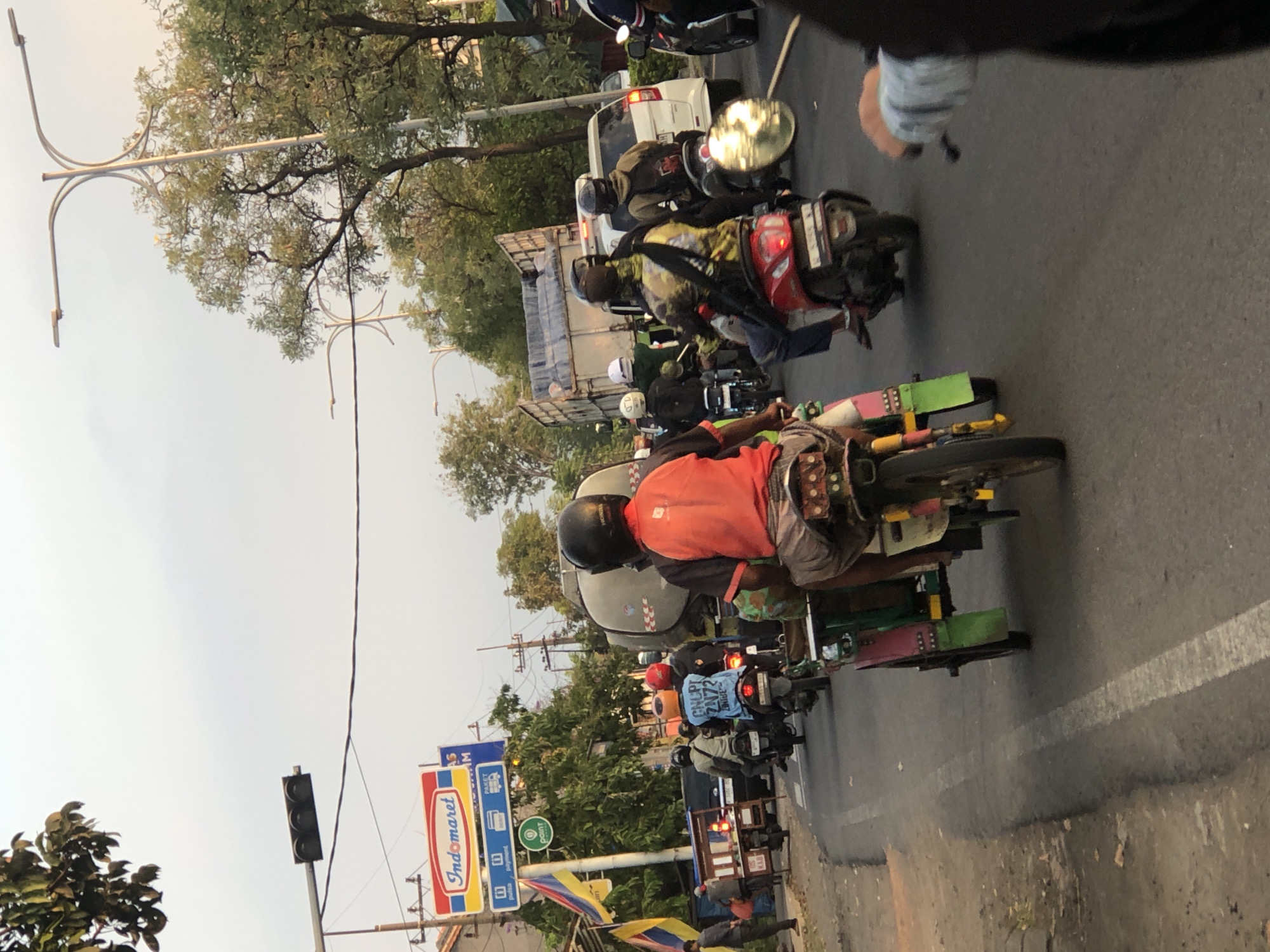 In Kludan, Indonesia, a street scene unfolds with moderate traffic. Several motorcycles navigate the asphalt road, some carrying single riders, while others transport passengers or cargo. One motorcycle is attached to a small, multi-colored cart. Among the riders, some wear helmets for protection, ranging in color from black to red and white. In the background, a white car and a large truck with a grey cargo bed are also present. The environment features trees lining the road and utility poles with wires overhead. A traffic light is visible, suggesting an intersection or a controlled roadway. A prominent sign for "Indomaret" stands out, indicating a commercial establishment nearby. The sky appears overcast, with a soft, diffused light, hinting at either early morning or late afternoon.