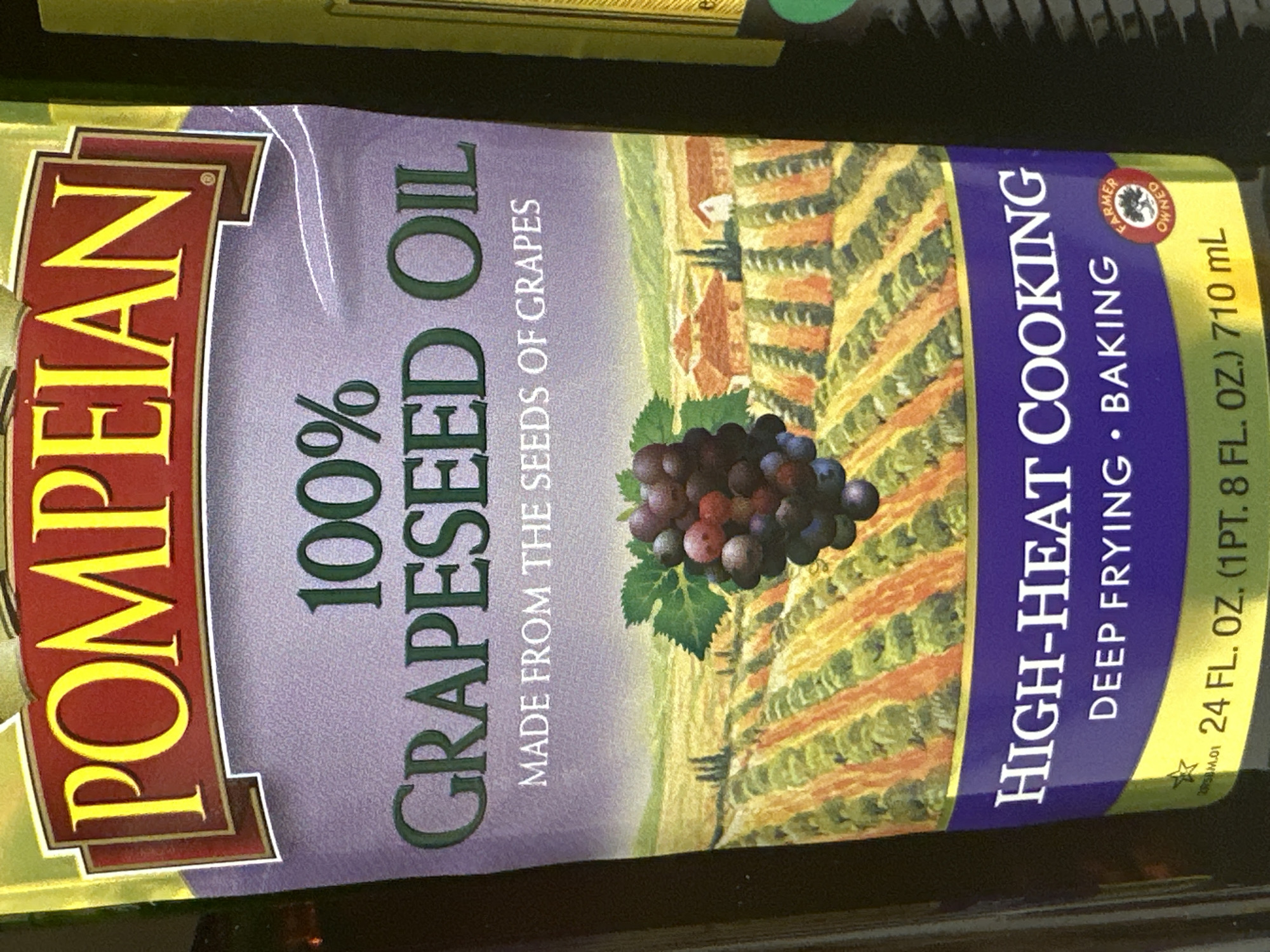 The image is a close-up of a bottle of Pompeian Grapeseed Oil, with a landscape scene featuring a vineyard and houses. The label prominently displays "POMPEIAN 100% GRAPESEED OIL MADE FROM THE SEEDS OF GRAPES". Below the vineyard image, it reads "HIGH-HEAT COOKING DEEP FRYING • BAKING" and includes a "FARMER OWNED" logo. The bottom of the label shows "24 FL. OZ. (1 PT. 8 FL. OZ.) 710 mL". The bottle appears to be on display in a retail environment, likely a grocery store, given the lighting and framing. The background is dark, suggesting it is either shelf packaging or a dimly lit aisle. The specific location of Camp Hill, United States of America, is not directly evident from the image itself but might be inferred from external context not provided.