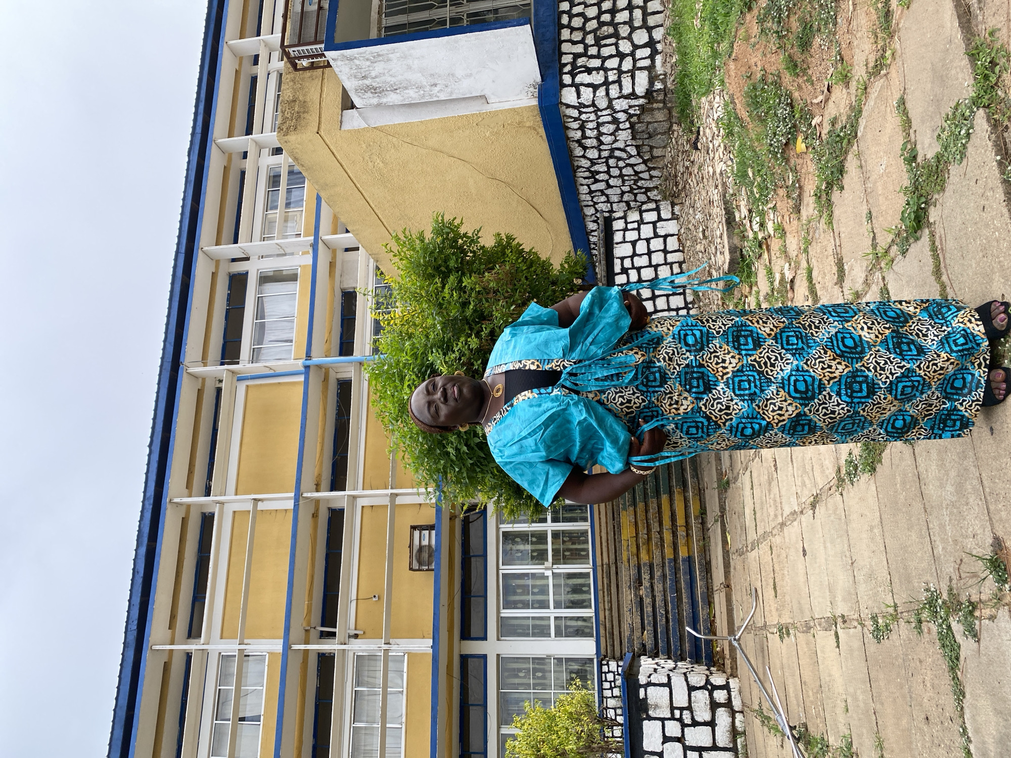 The image features a woman standing outdoors, facing the viewer with a gentle smile, in Bukuru, Nigeria. She is positioned slightly to the right of the frame, wearing a vibrant attire that includes a blue, short-sleeved top over a black garment, and a long skirt or dress with a distinct geometric pattern in blue, black, and tan. She has a dark head covering, gold-colored earrings, a necklace, and wears sandals.
Behind the woman is a multi-story building, primarily painted in a light yellow hue with prominent blue trim along its roofline and around its numerous windows. The windows are large, some featuring white frames, giving the building a structured, possibly institutional, appearance. Green foliage and bushes are visible behind and to the side of the woman, between her and the building.
The ground she stands on is a concrete path, showing signs of wear with cracks and small patches of green weeds growing through them. To the lower right, a low wall or structure with a black and white block pattern is visible. The sky above is overcast and light grey, suggesting daytime under cloudy conditions. There is a small metal stand or pole on the ground near her feet. No visible text is present in the image.