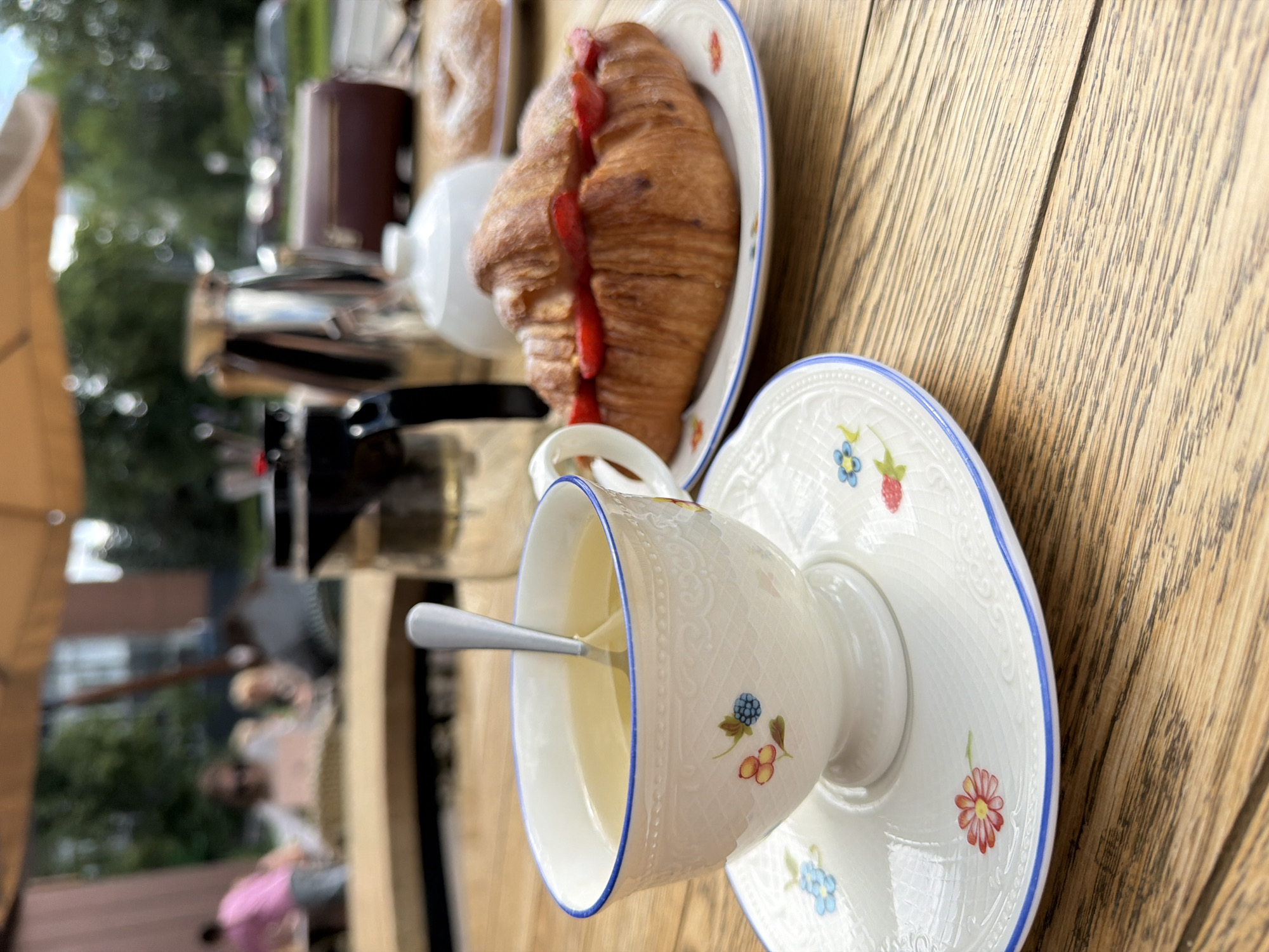 A close-up shot captures a table setting at an outdoor cafe, likely in Izhevsk, Russia, given the context. The scene features a delicate white teacup with blue trim and floral patterns, filled with a pale yellow liquid and a silver spoon submerged within. The teacup rests on a matching saucer, also adorned with similar floral designs. To the right, a golden-brown croissant, sliced and filled with vibrant red strawberry slices, sits on another plate. In the background, a tall glass French press and a metallic thermos are visible. Further back, out of focus, are other patrons and the blurred greenery of trees, suggesting a pleasant, casual outdoor dining environment. The wooden table surface is rustic and weathered.