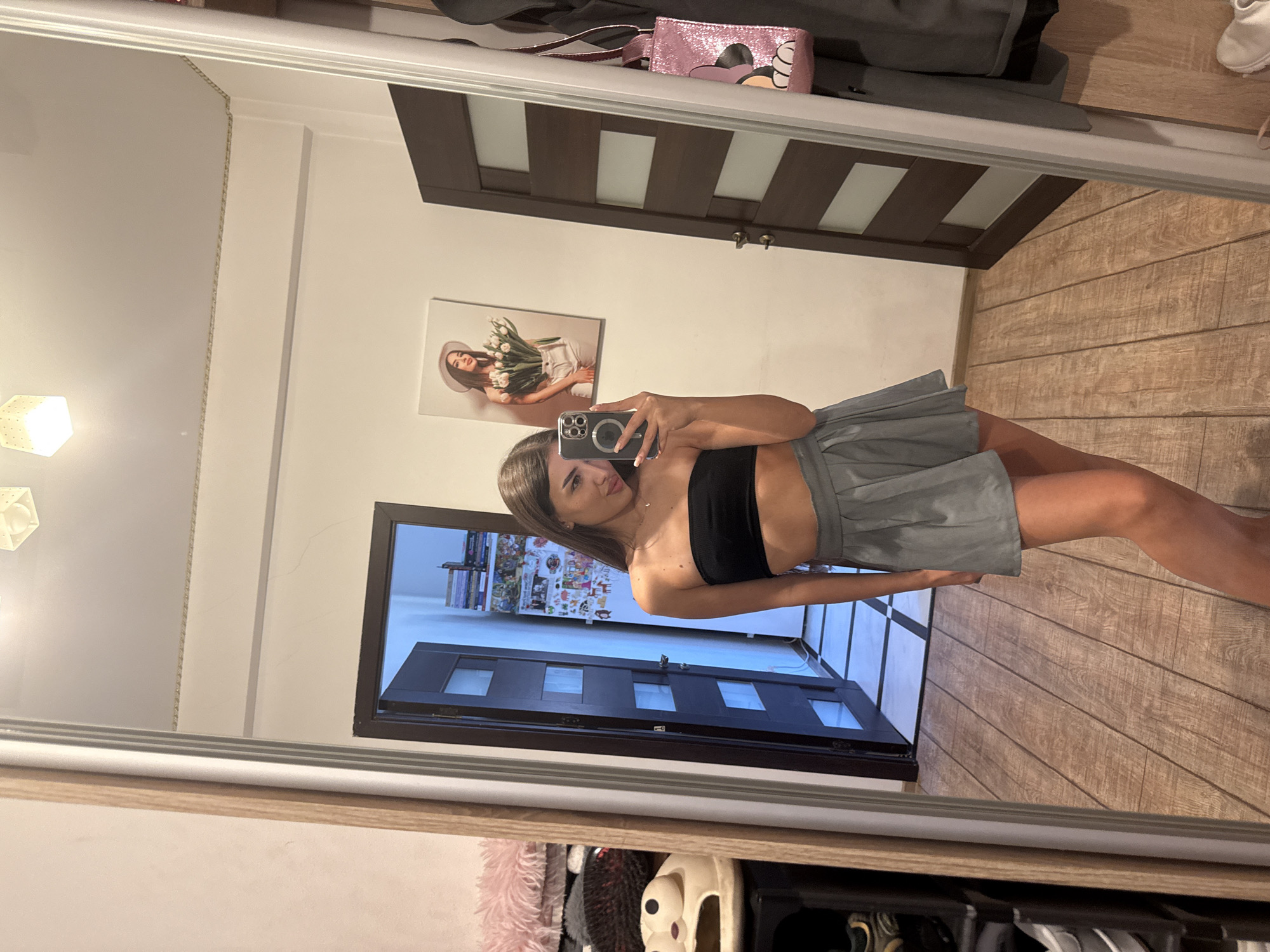 A young woman with long, brown hair is taking a mirror selfie in a room. She is wearing a black strapless top and a grey pleated skirt. She is holding a smartphone in her right hand, capturing her reflection. Behind her, a large mirror reflects the room, showing a doorway with a partially open door, a painting on the wall depicting a woman with flowers, and other decorative elements. The room has wooden flooring and cream-colored walls. To the left of the mirror, there is a shelf unit with various items, including what appears to be stuffed animals or plush toys. To the right of the mirror, a dark brown door is visible, and a pink glittery bag with a Mickey Mouse design hangs on a hook. The overall setting appears to be a bedroom or dressing area within a residential apartment. There is no indication of the specific location in the city of Chișinău, Moldova.