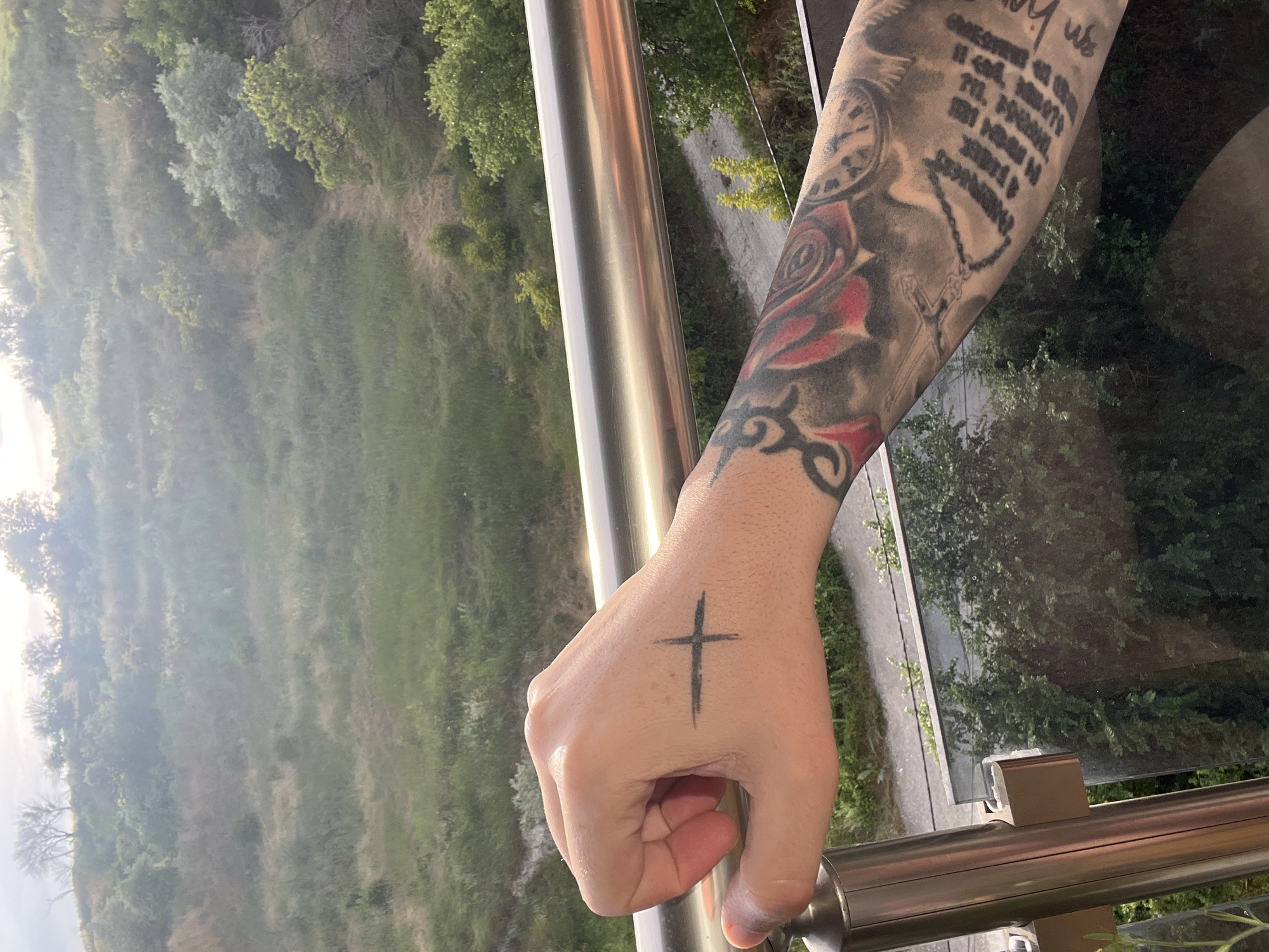 A person's tattooed arm and hand rest on a railing overlooking a natural landscape with a gradient of green vegetation. The hand, positioned over the railing, displays a simple, dark cross tattoo on the back. The forearm features extensive, dark-ink tattoos, including a prominent red rose design and lettering. The railing is made of shiny metal. The background depicts a hilly, overgrown area with a path winding through it, under a bright, hazy sky. This scenery is consistent with the coastal hills found in the Vinitsa district of Varna, Bulgaria.