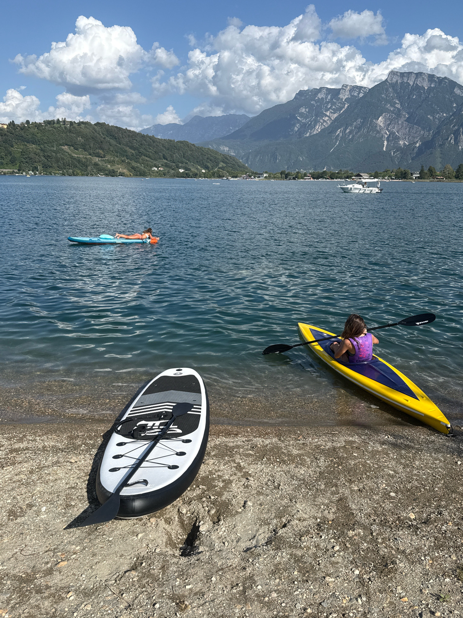 The image captures a serene summer day at Calceranica al Lago, Italy, with a focus on water activities. In the foreground, a stand-up paddleboard lies on the pebbly shore, its black and white deck contrasting with the water. To the right, a yellow and blue kayak is partially submerged at the water's edge, with a young person wearing a purple life vest seated inside, holding a paddle. Further out on the calm, blue water, another person is lying on a bright blue inflatable raft. The background features a lush green hillside dotted with buildings, leading up to imposing, rocky mountains under a sky filled with dramatic white clouds. A white boat is visible in the distance on the lake. The overall scene is one of peaceful recreation in a beautiful natural setting.